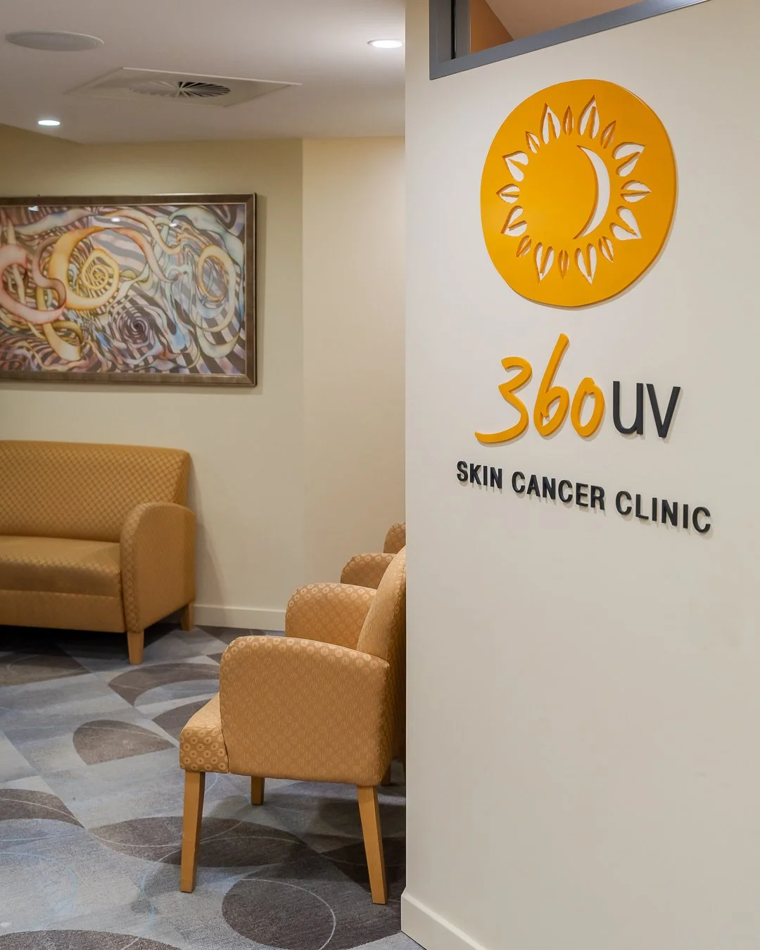Interior of a skin cancer clinic with a sign reading "360 UV Skin Cancer Clinic" on a white wall. There are two beige chairs and a beige sofa, with a colorful abstract painting hanging on the wall in the background.