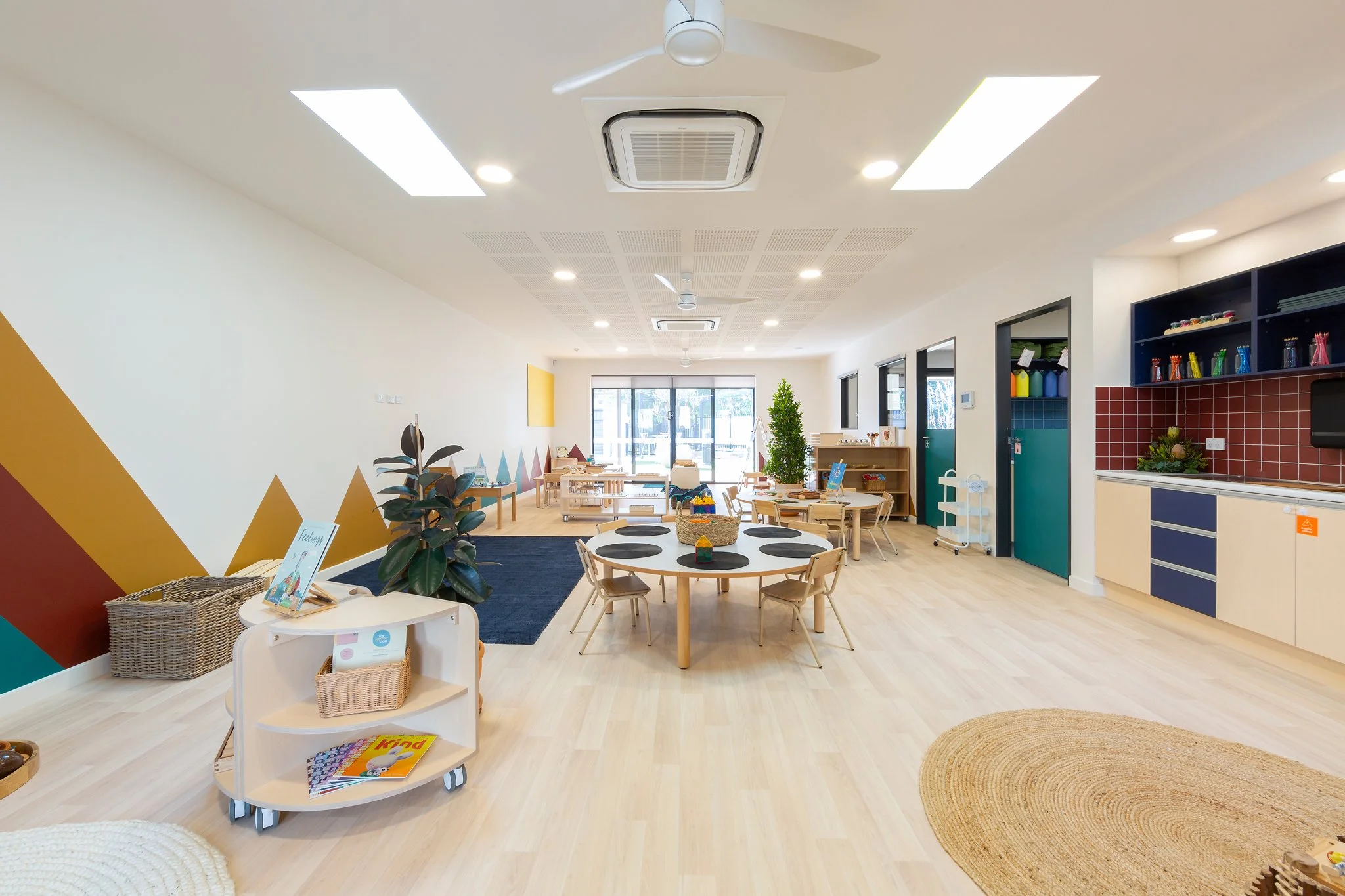 A brightly lit classroom with wooden furniture, a colorful geometric wall mural, and a variety of educational toys and books, including a round table with chairs, a small white bookshelf with toys, and a bookshelf with art supplies and decorative pla