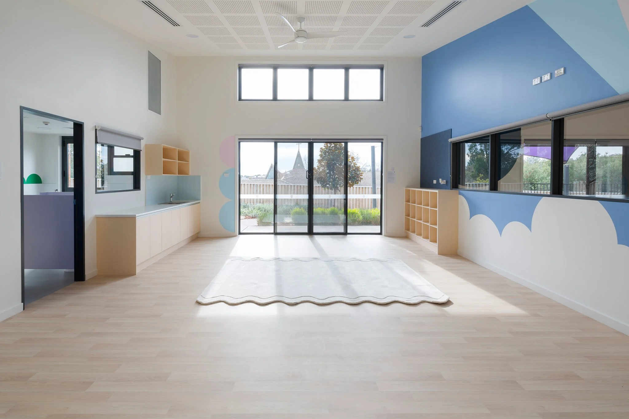 A spacious room with light-colored wooden flooring, white and blue walls with abstract patterns, large windows allowing natural light, a ceiling fan, and built-in shelving.