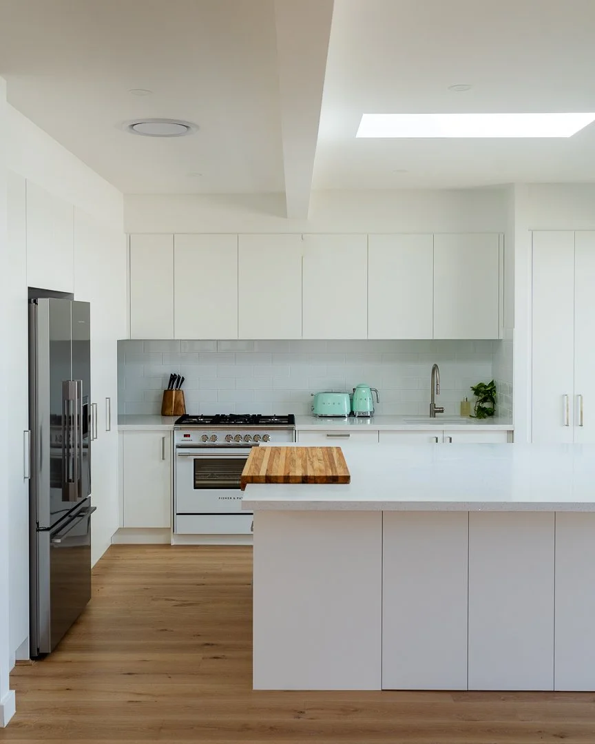 Modern white kitchen with stainless steel refrigerator, stove, mint green toaster and kettle, white cabinetry, wooden cutting board, wood flooring, and a window letting in natural light.