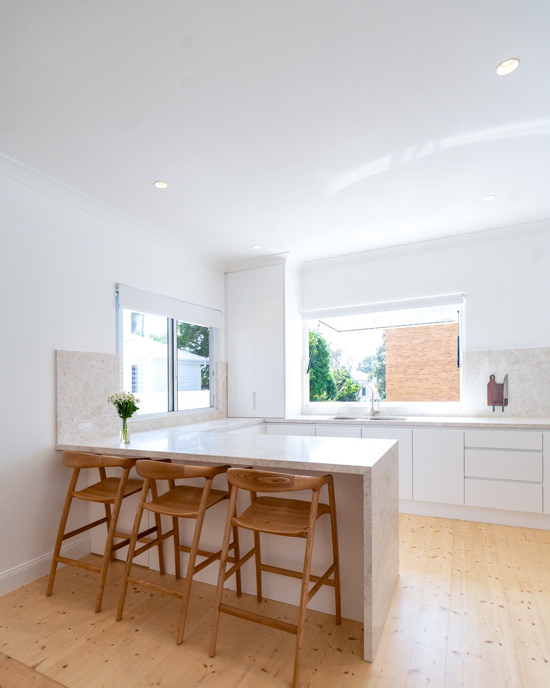 Modern white kitchen with a marble countertop island, three wooden bar stools, and large windows letting in natural light, overlooking trees and a brick building.