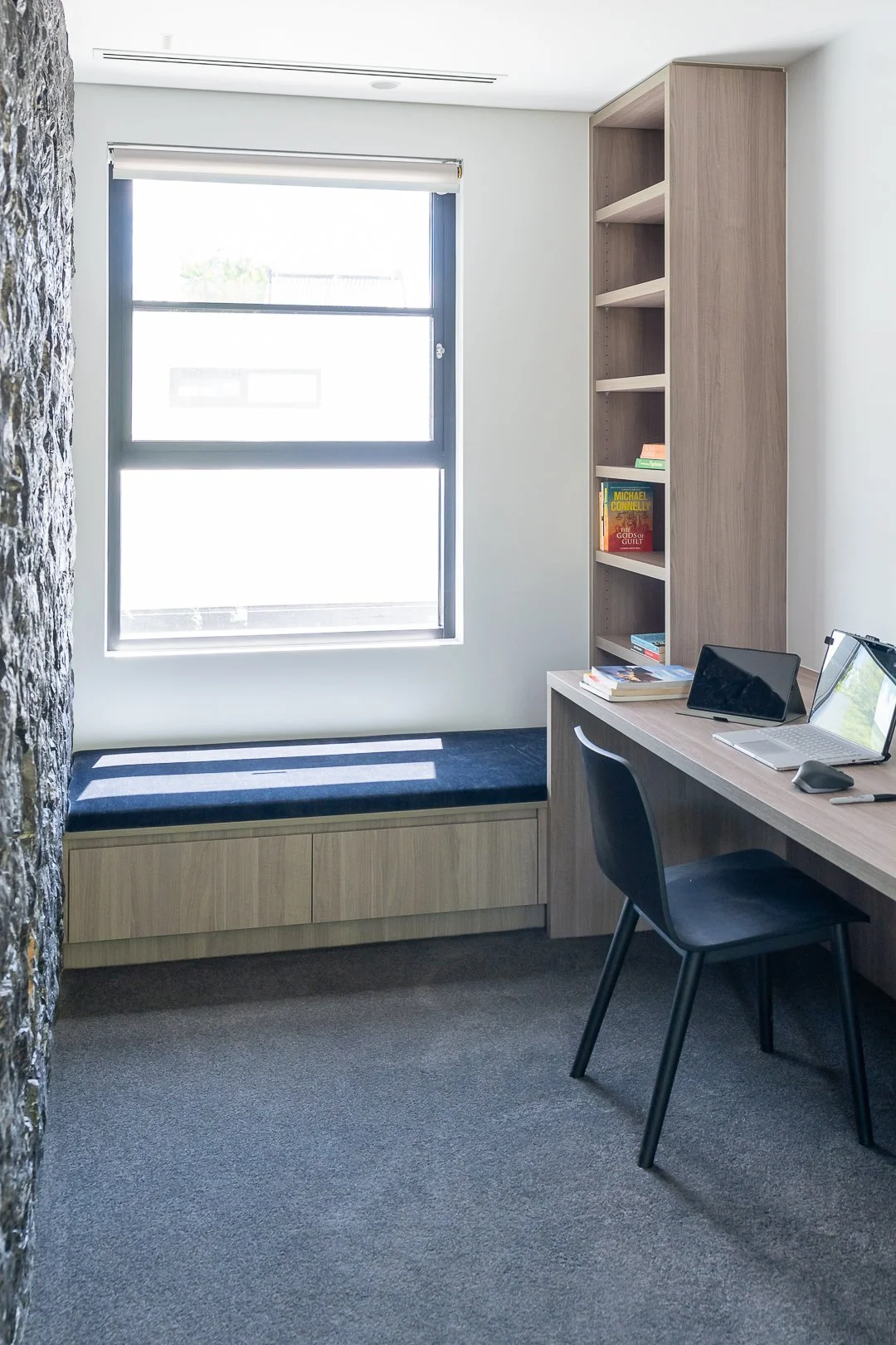 A small, modern home office with a window seat, bookshelf, and desk. The window lets in natural light, and there are a few books on the shelf and desk. The room has neutral-colored walls, a textured wall on one side, and gray carpet flooring.