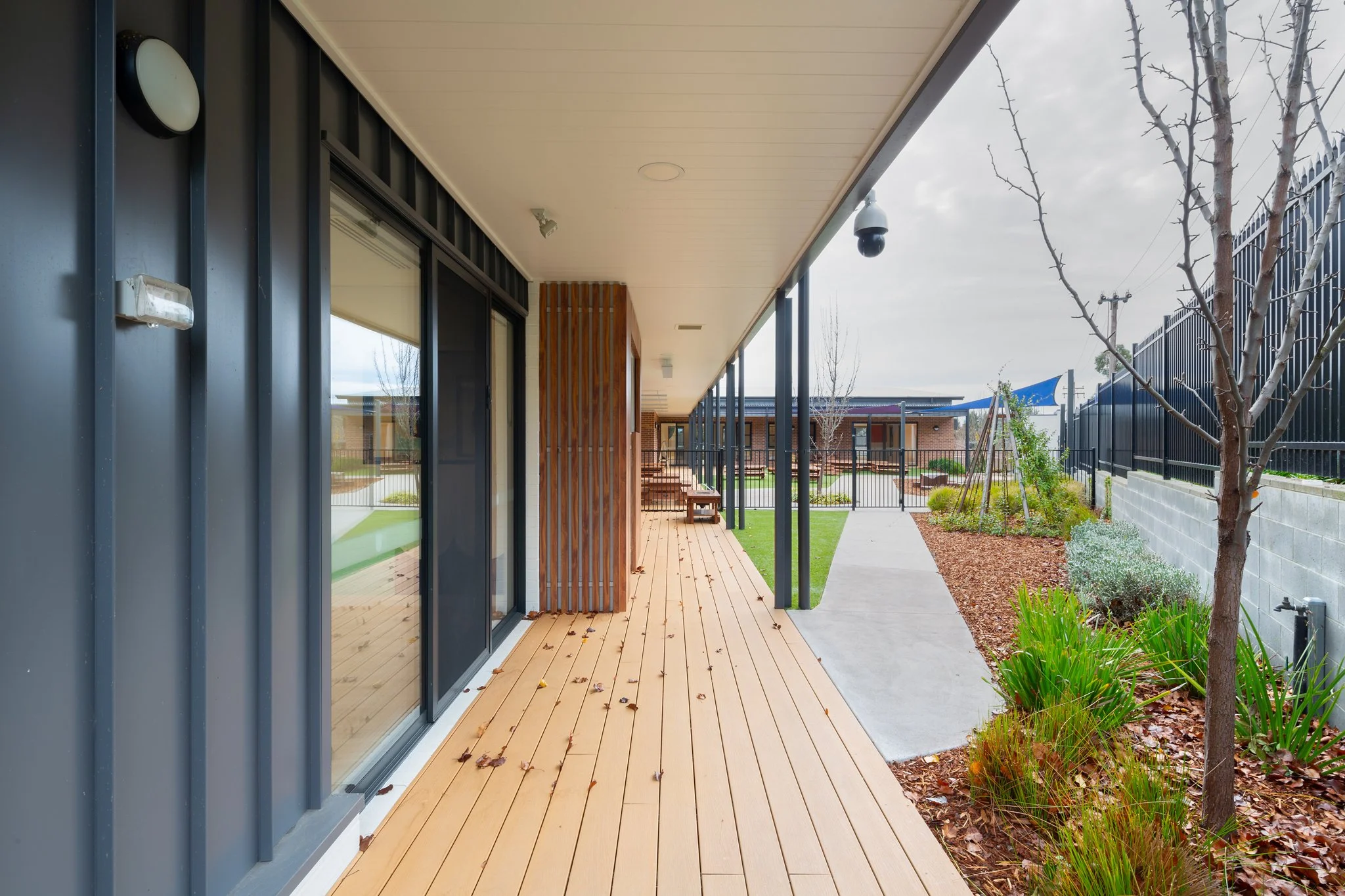 A modern house's covered porch with a wooden deck, large sliding glass doors, and a landscaped yard with small trees and plants.