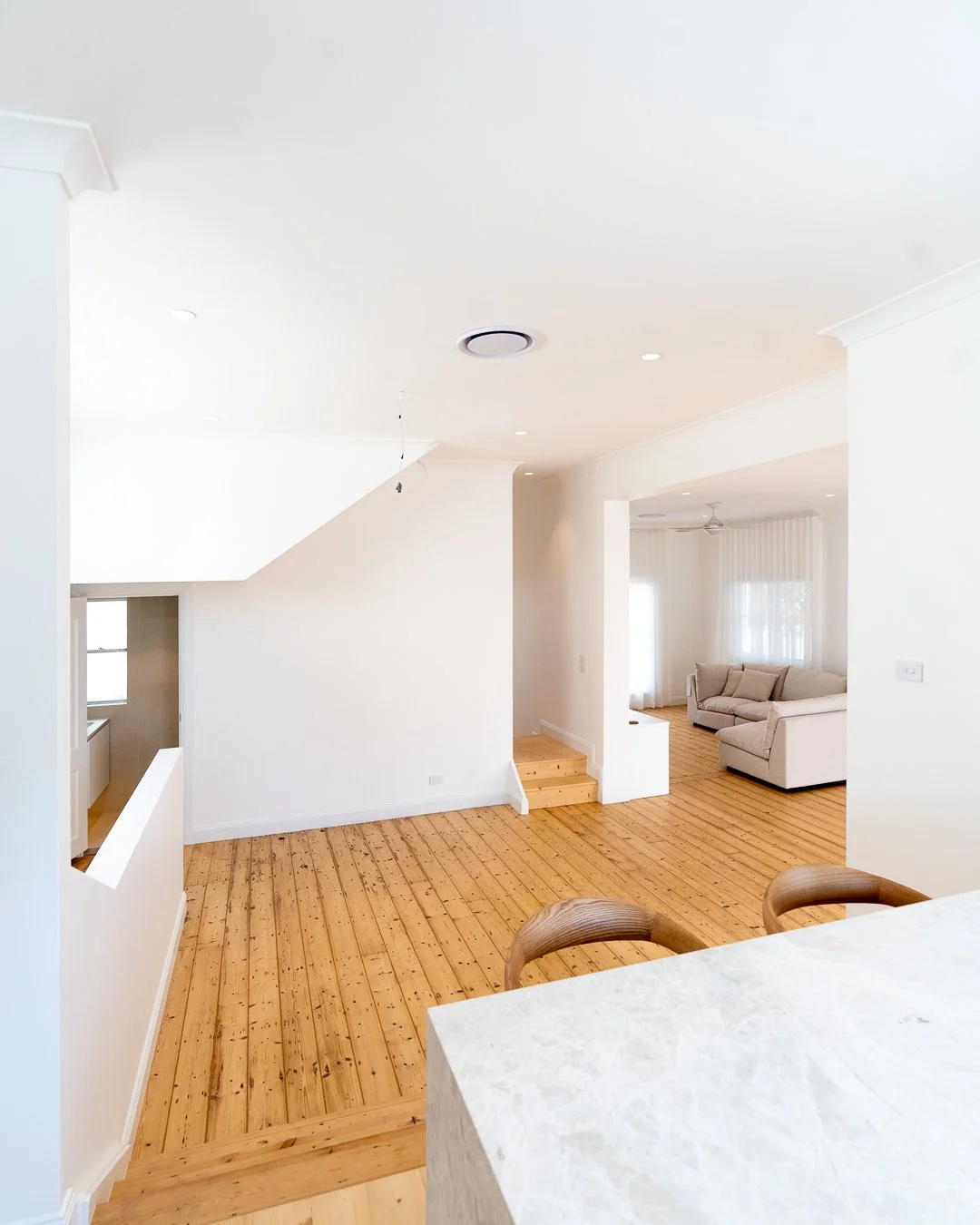Bright, open-concept living area with white walls, wooden flooring, and beige furniture including a sofa and chairs.