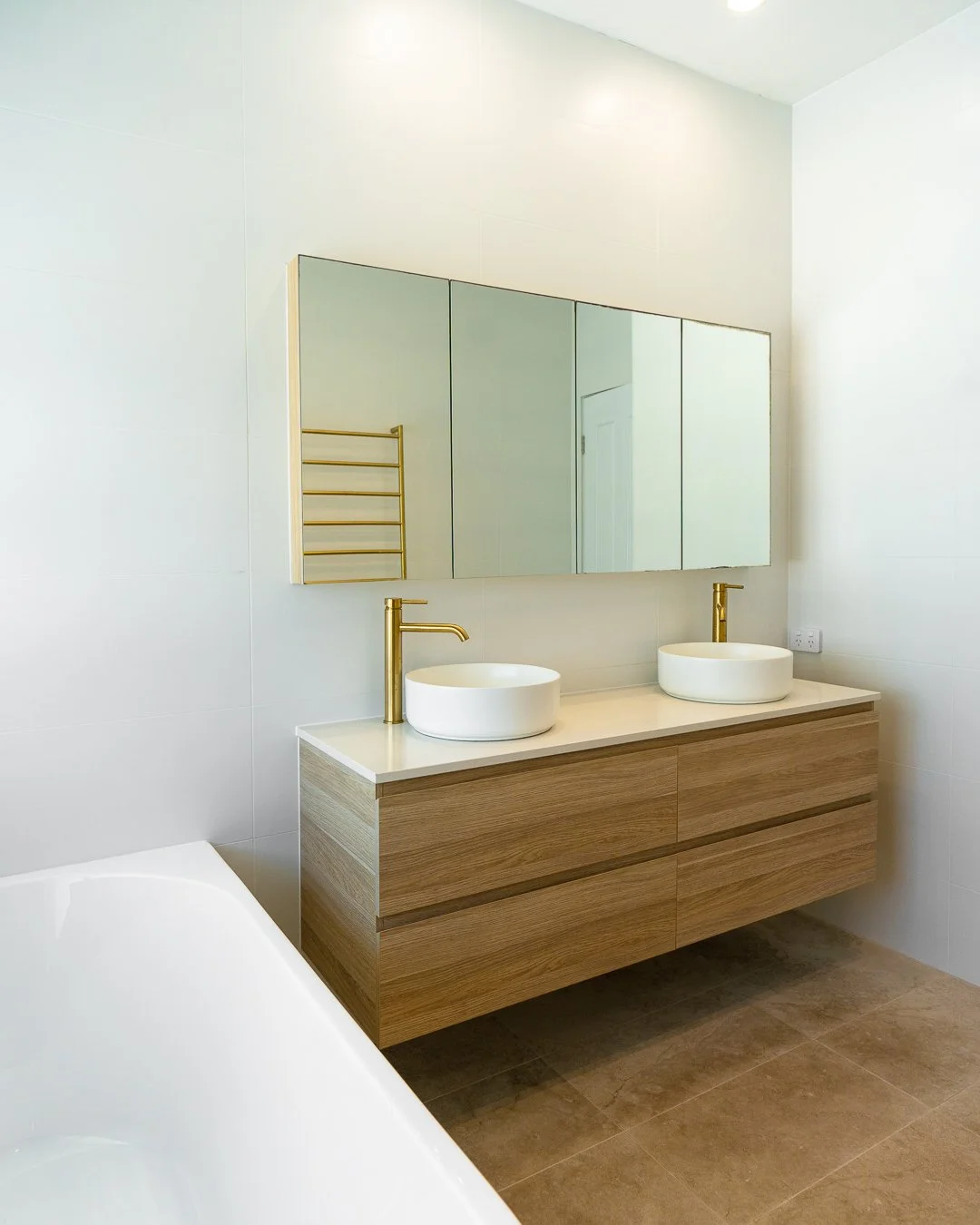 Modern bathroom with a double vanity, white vessel sinks, gold faucets, a large mirror, a white bathtub, and wood cabinets, with beige tiled flooring.
