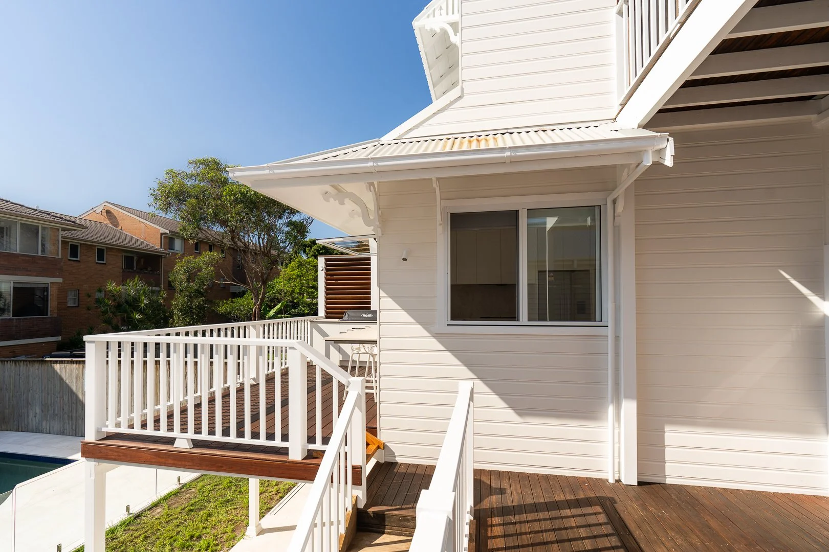 Exterior view of a white house with a wooden deck, white railing, and large window, surrounded by neighboring houses and trees under a clear blue sky.