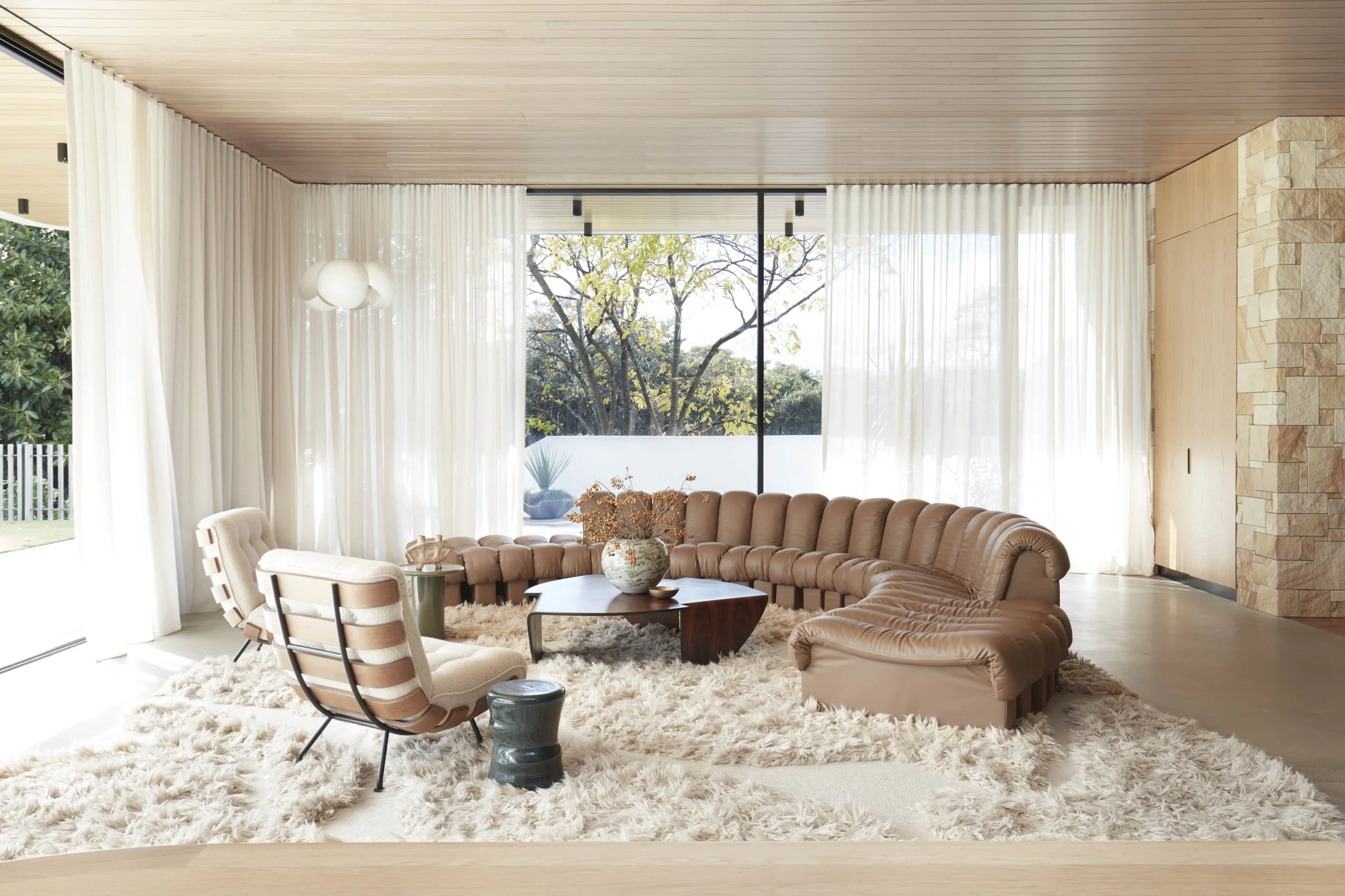 Modern living room with large windows, white curtains, beige sectional sofa, armchair, and a fluffy white rug.