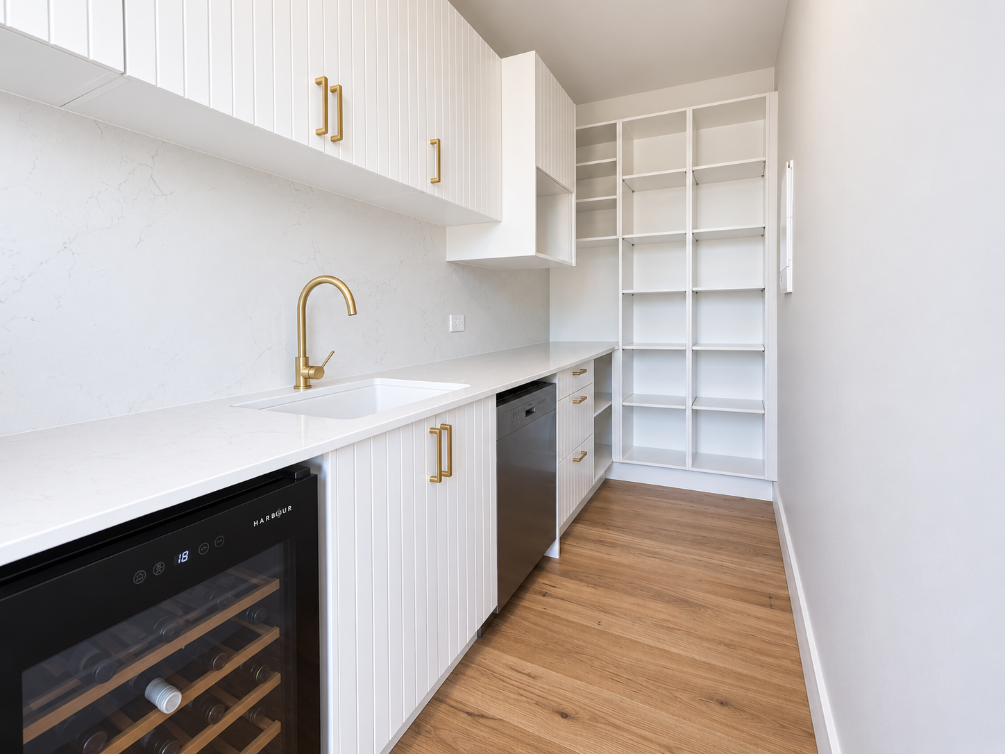 Modern laundry room with white cabinets, gold handles, a built-in wine cooler, a white countertop, gold faucet, and open shelving units, with hardwood flooring.