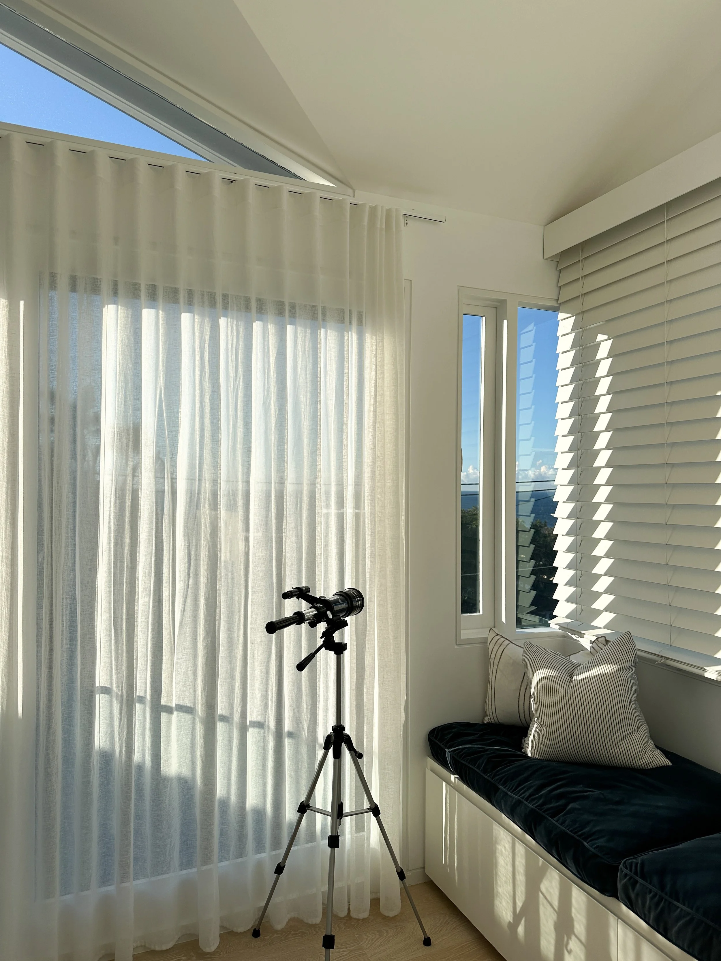 A room with large windows covered by sheer white curtains and adjustable horizontal blinds. There is a black telescope on a tripod, a bench with a black cushion, and pillows with striped covers, with a view of the blue sky and clouds outside.