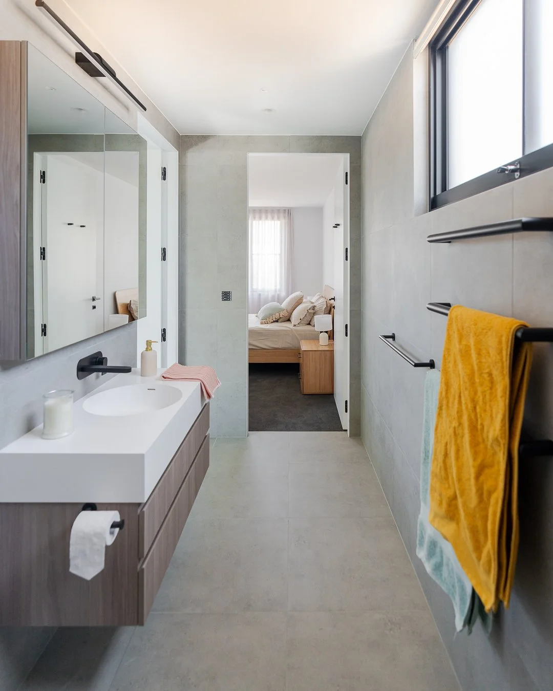 Modern bathroom with a vanity mirror, sink with soap dispenser, candles, and decorative towels. View into a bedroom with a bed and natural light from windows.