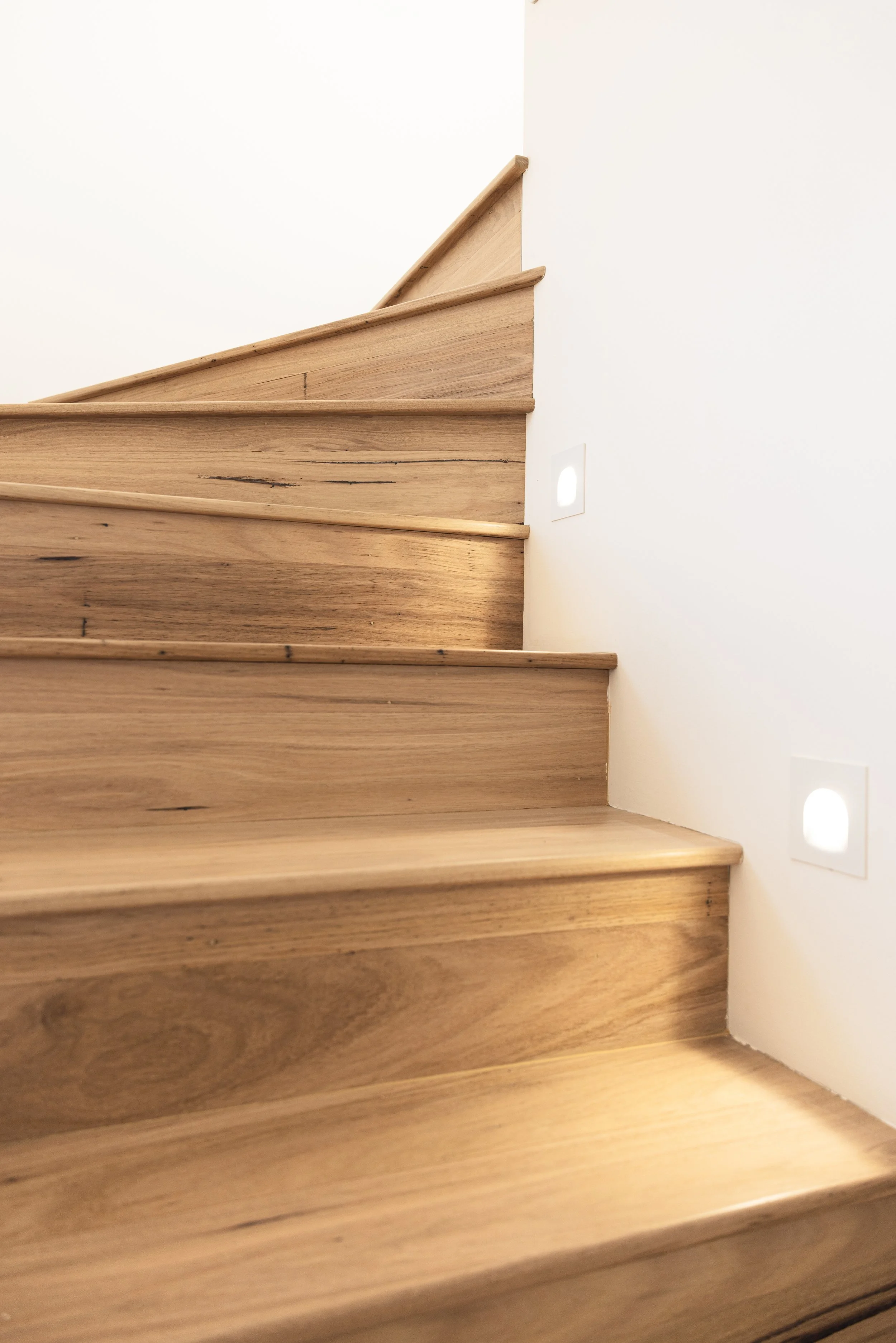 Close-up of a wooden staircase with light-colored wood and white wall with small wall lights.