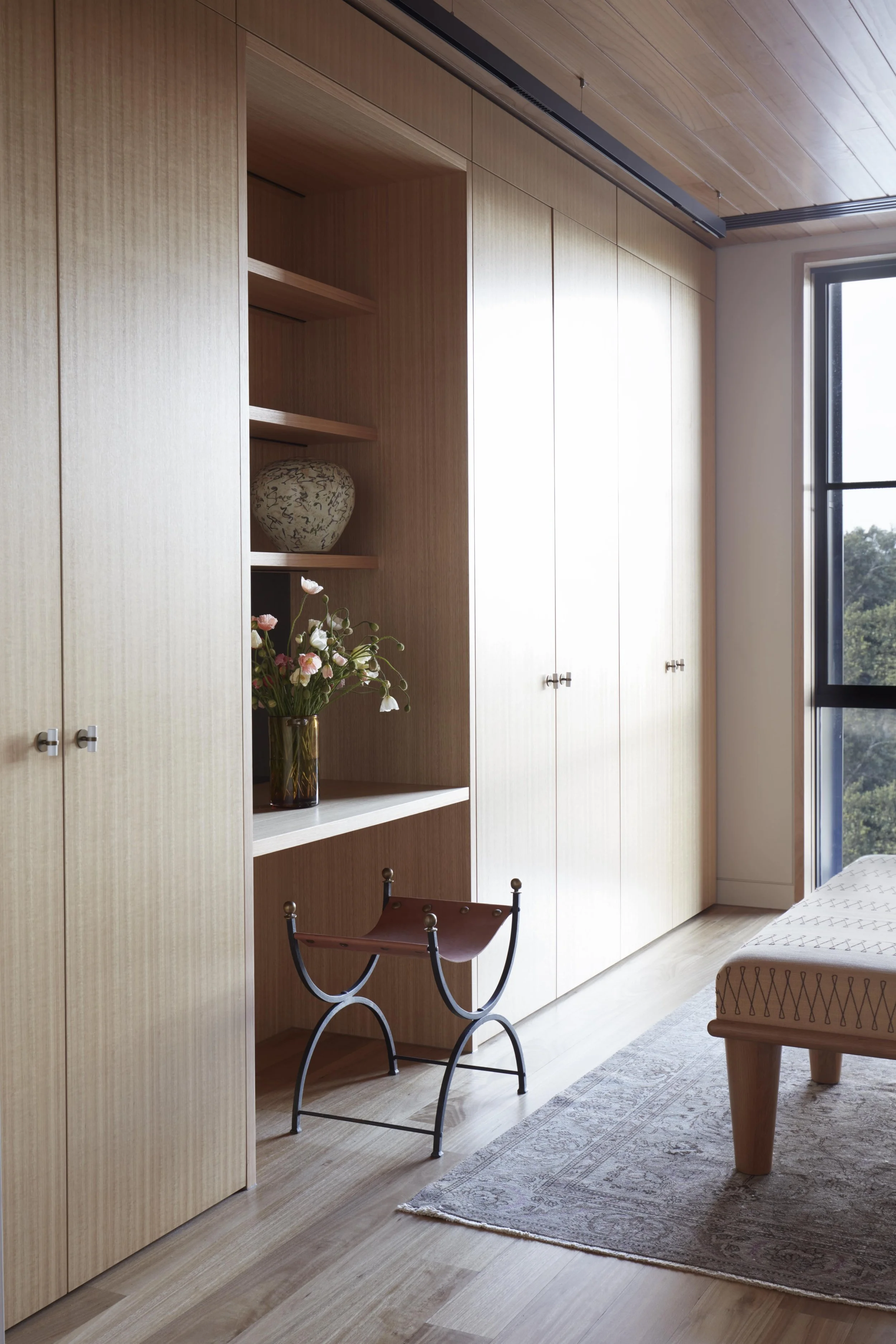 A room with wooden built-in cabinets, a bottle with flowers, a chair, a rug, and a window showing trees outside.
