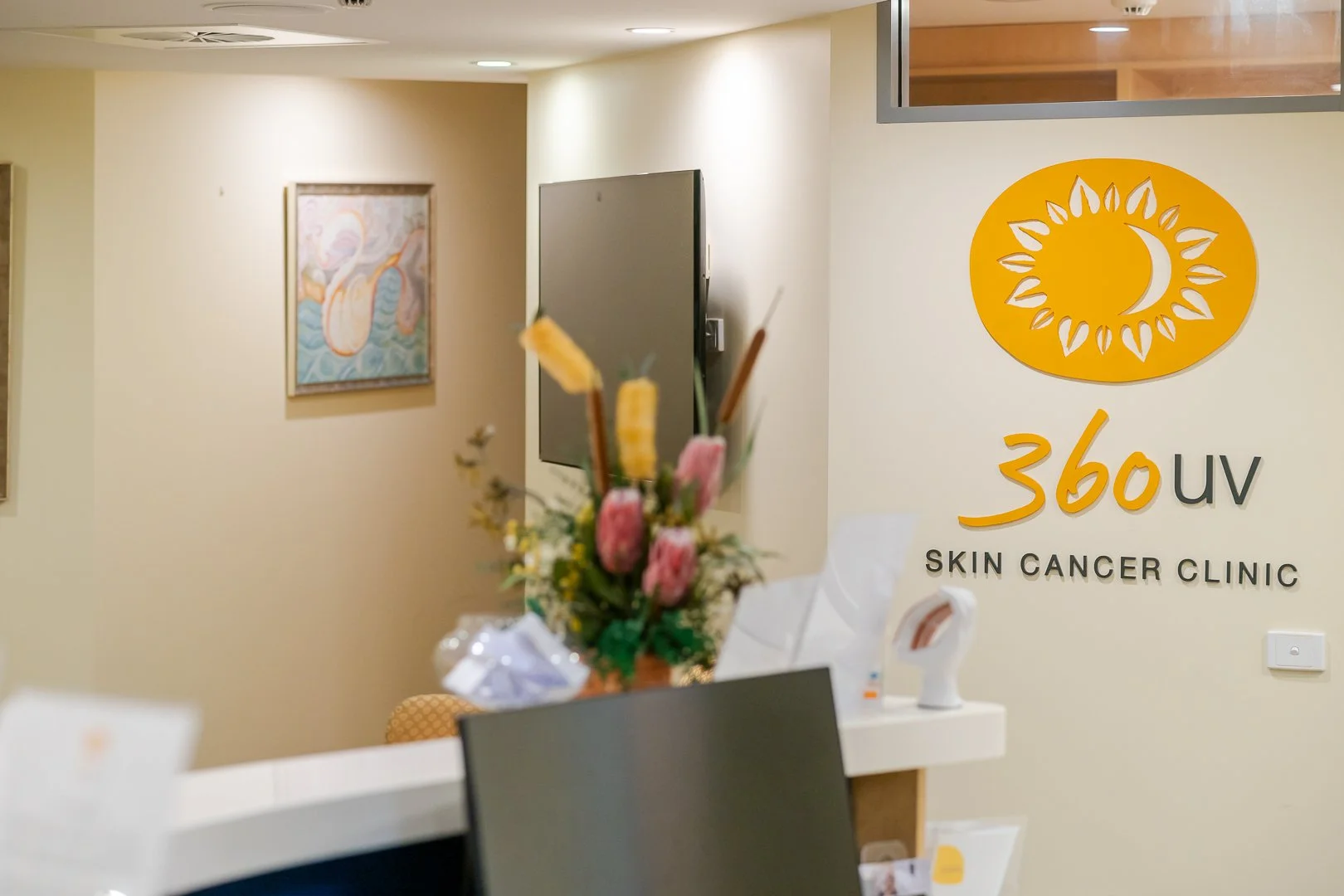 Reception area of a skin cancer clinic with a logo for 360 UV Skin Cancer Clinic on the wall, a flower arrangement on the counter, and framed artwork on the beige wall.