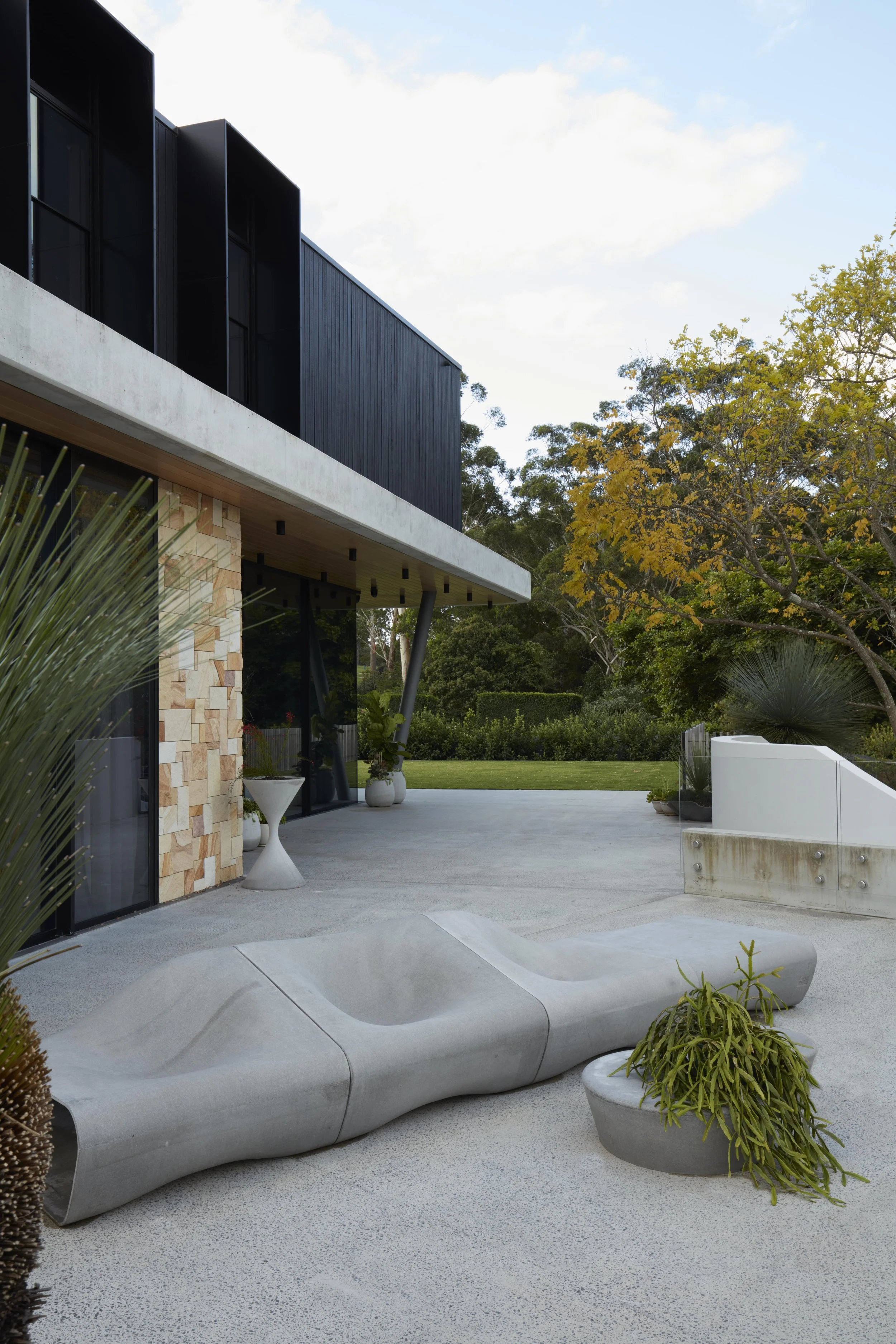 Modern building with black and stone exterior, outdoor seating, surrounded by trees and plants, with a blue sky.