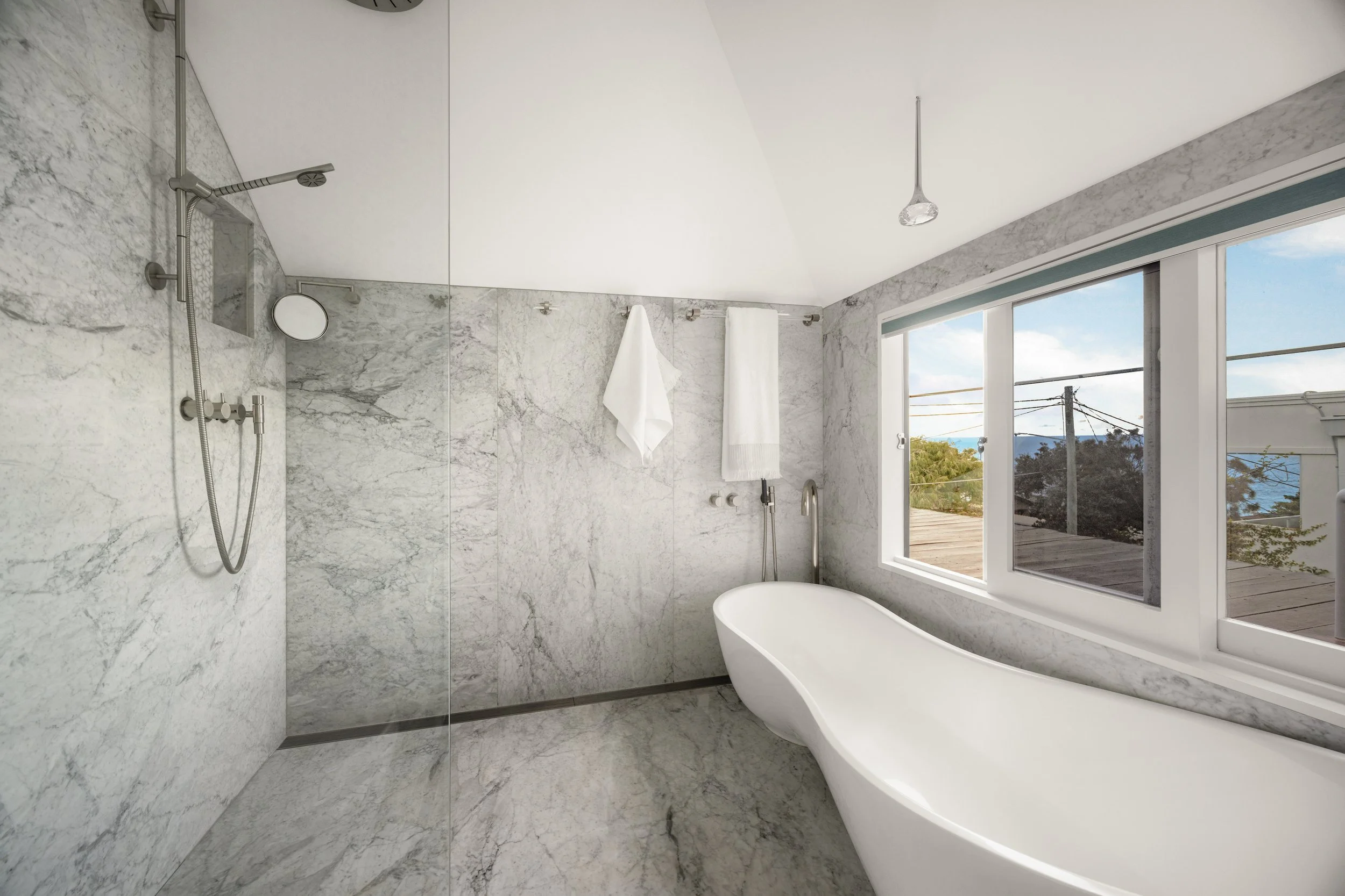 Modern bathroom with marble walls and flooring, a white free-standing bathtub, large window with sky view, and a ceiling shower head.