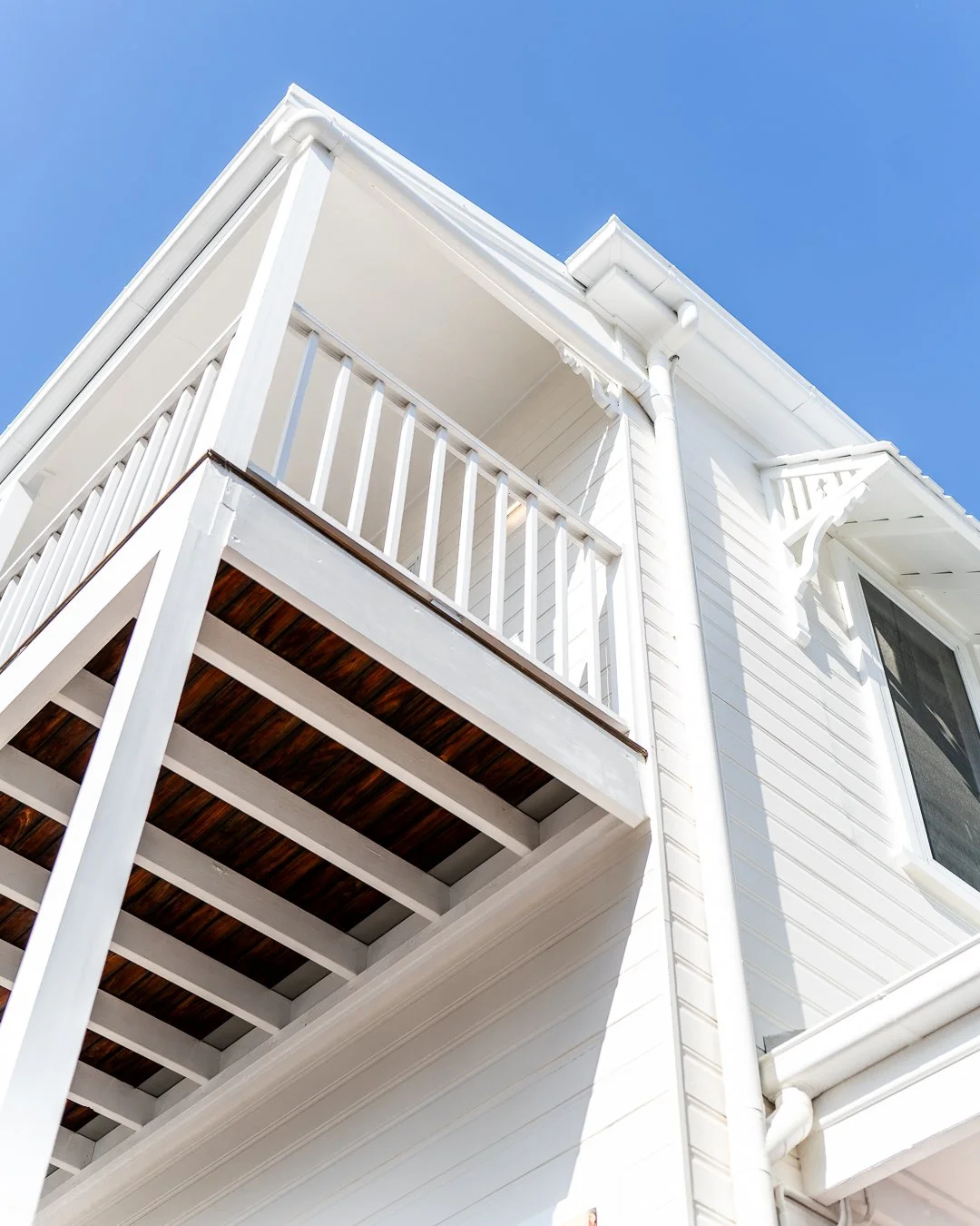 View of a white house exterior corner with balcony, wooden floor, and blue sky.