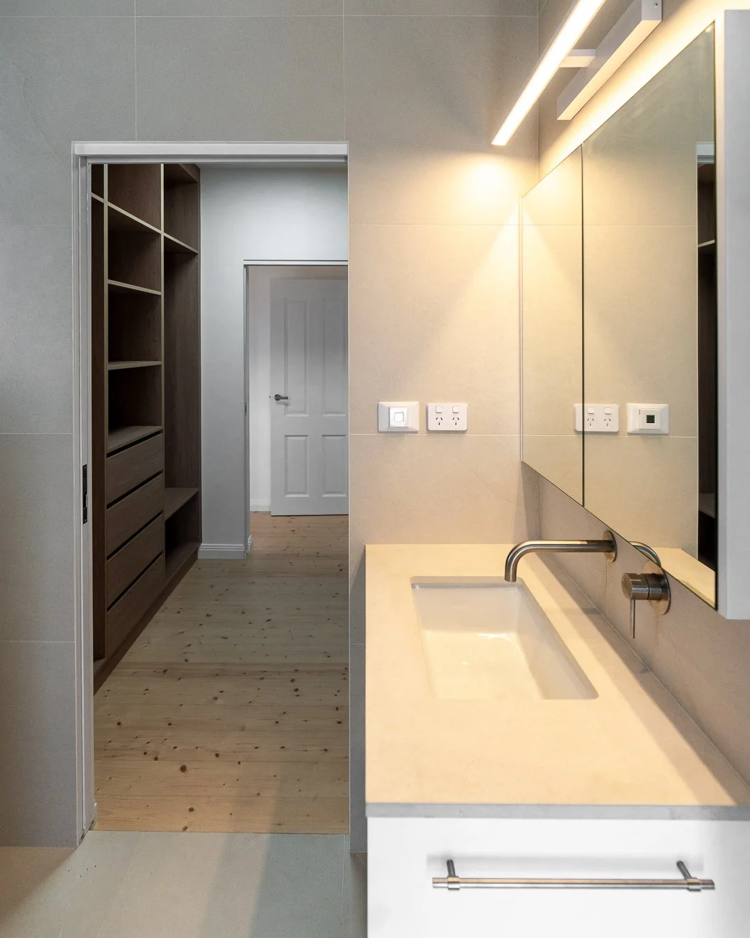 Modern bathroom with beige walls, a large mirror, and a sink with a brass faucet. There are electrical outlets on the wall and a towel bar beneath the sink. A doorway leads to a room with wooden flooring and built-in shelves or closet space.