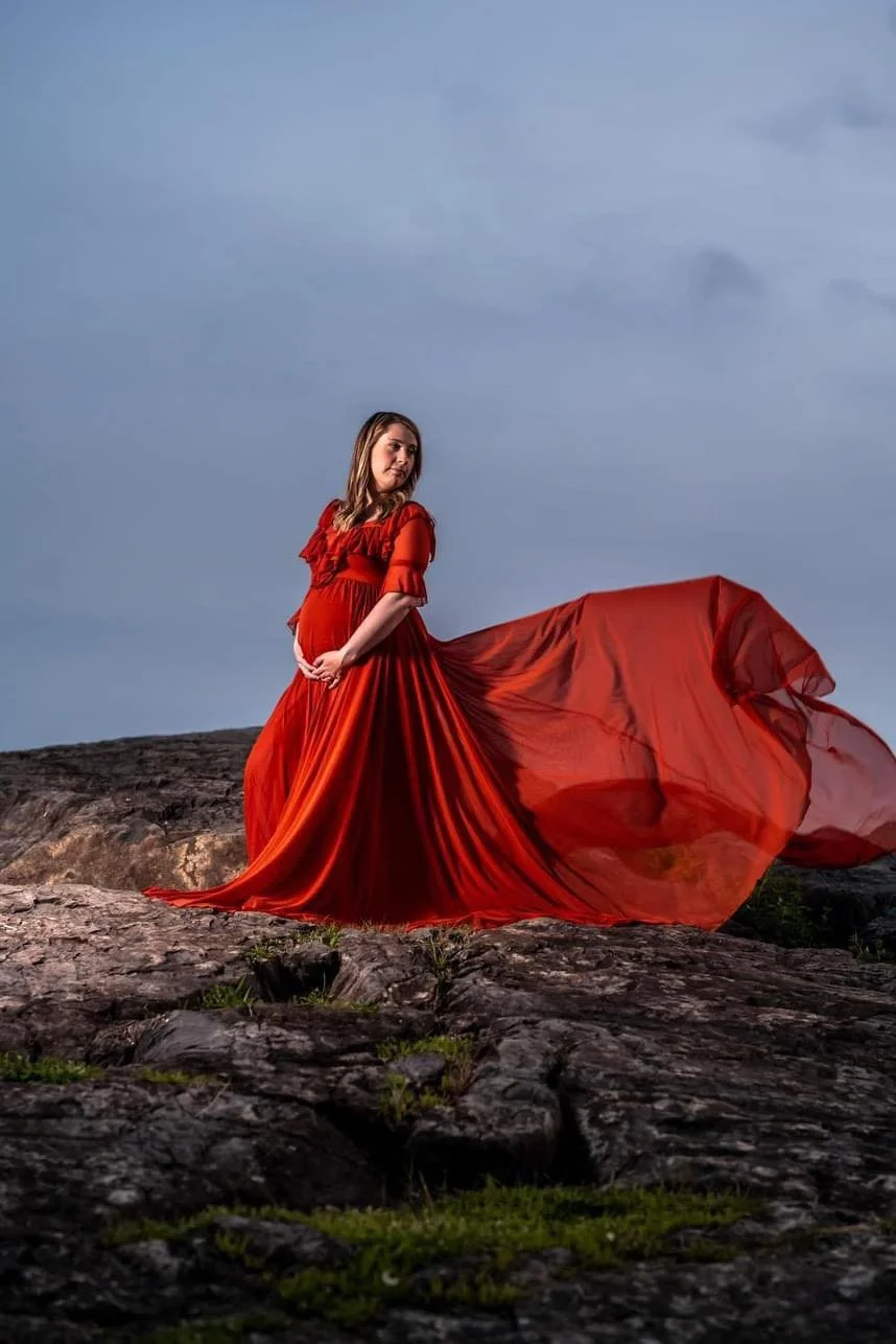 A pregnant woman in a flowing red dress standing on a rocky terrain outdoors with a cloudy sky in the background.