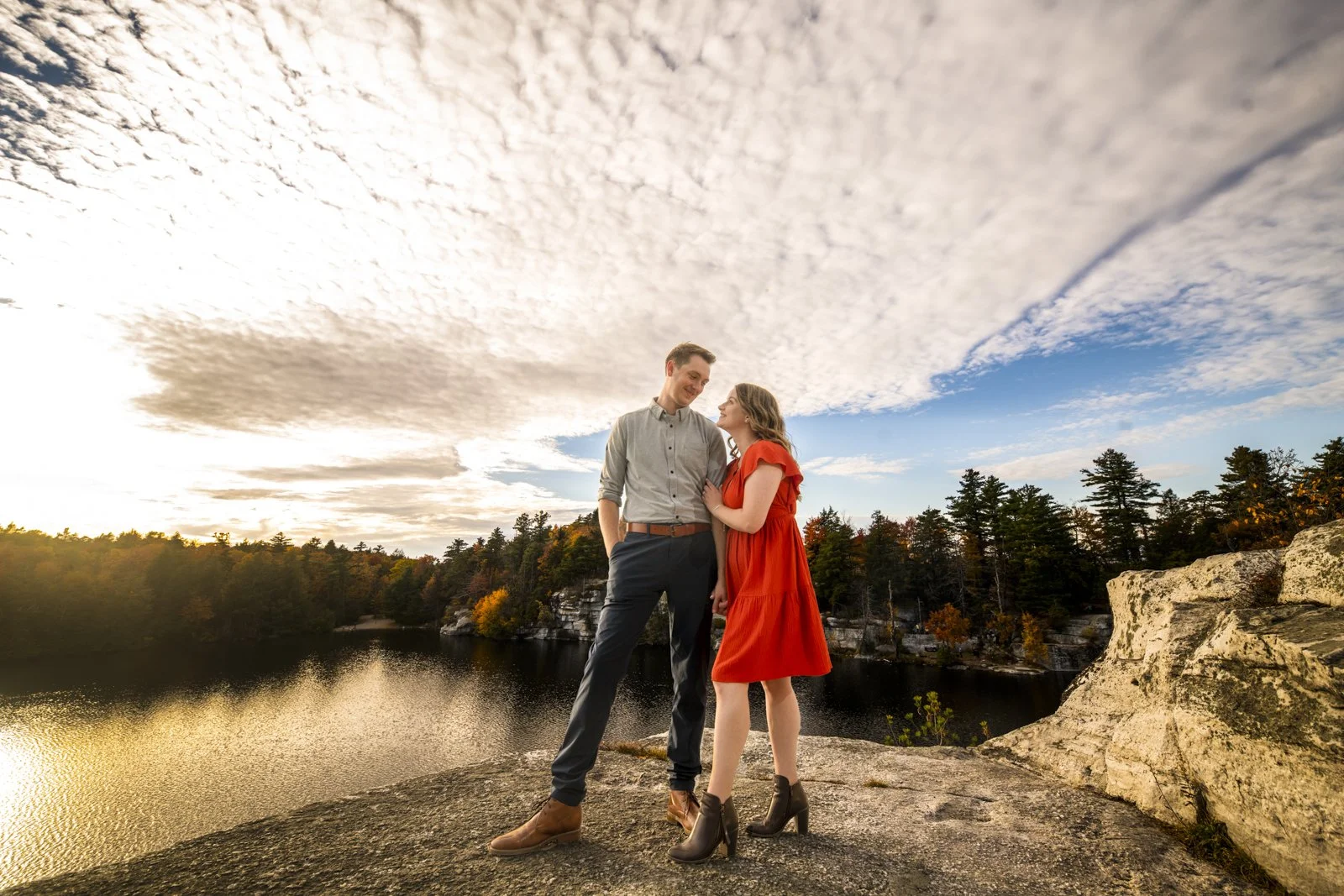 A smiling couple standing on a rocky ledge near a lake, with trees and an overcast sky in the background.