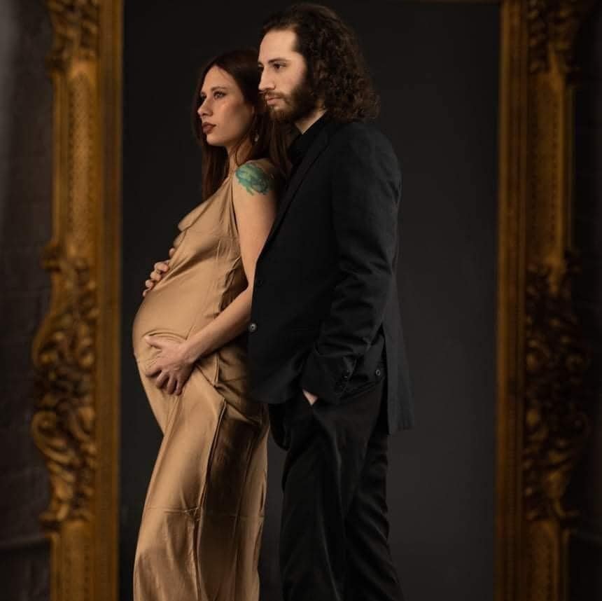 A pregnant woman with long hair and a green tattoo on her shoulder stands beside a man with curly hair and a beard, both in formal black clothing, looking thoughtfully into a mirror with a dark background and ornate gold frame.