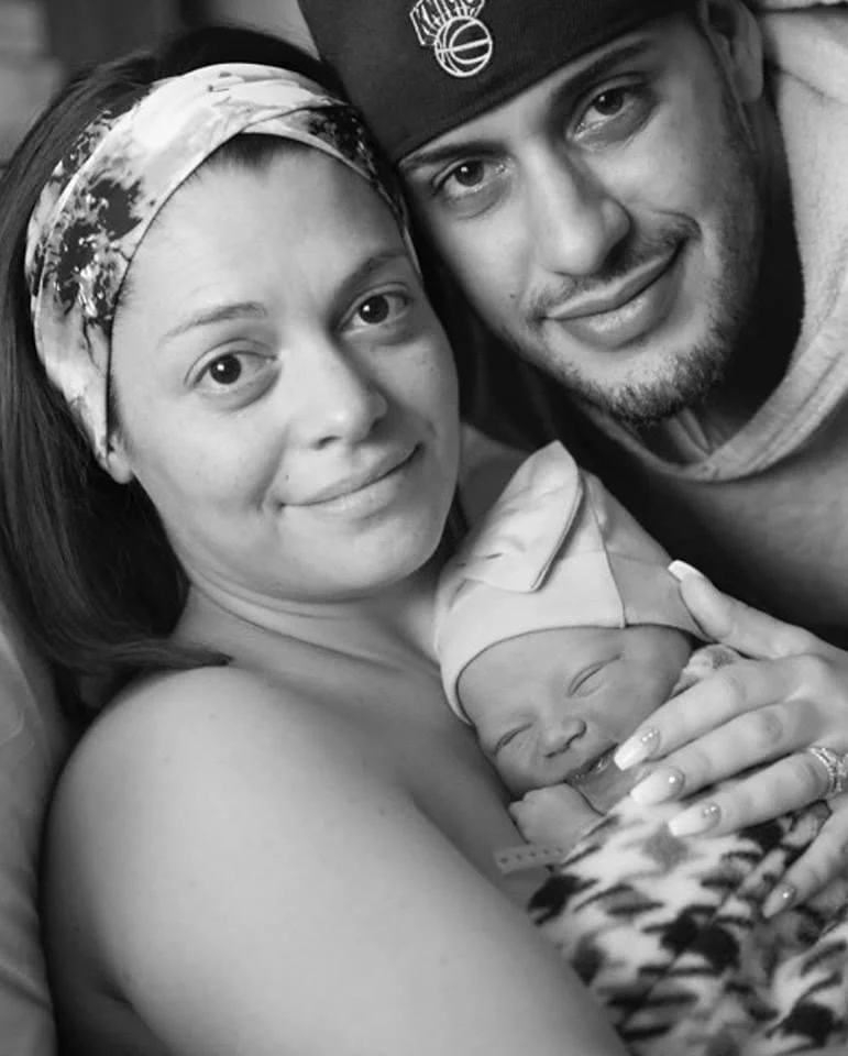 A woman, a man, and a newborn baby are lying close together; the woman is holding the baby, who is wearing a hat and blanket, and the man is leaning in beside them. The photo is in black and white.