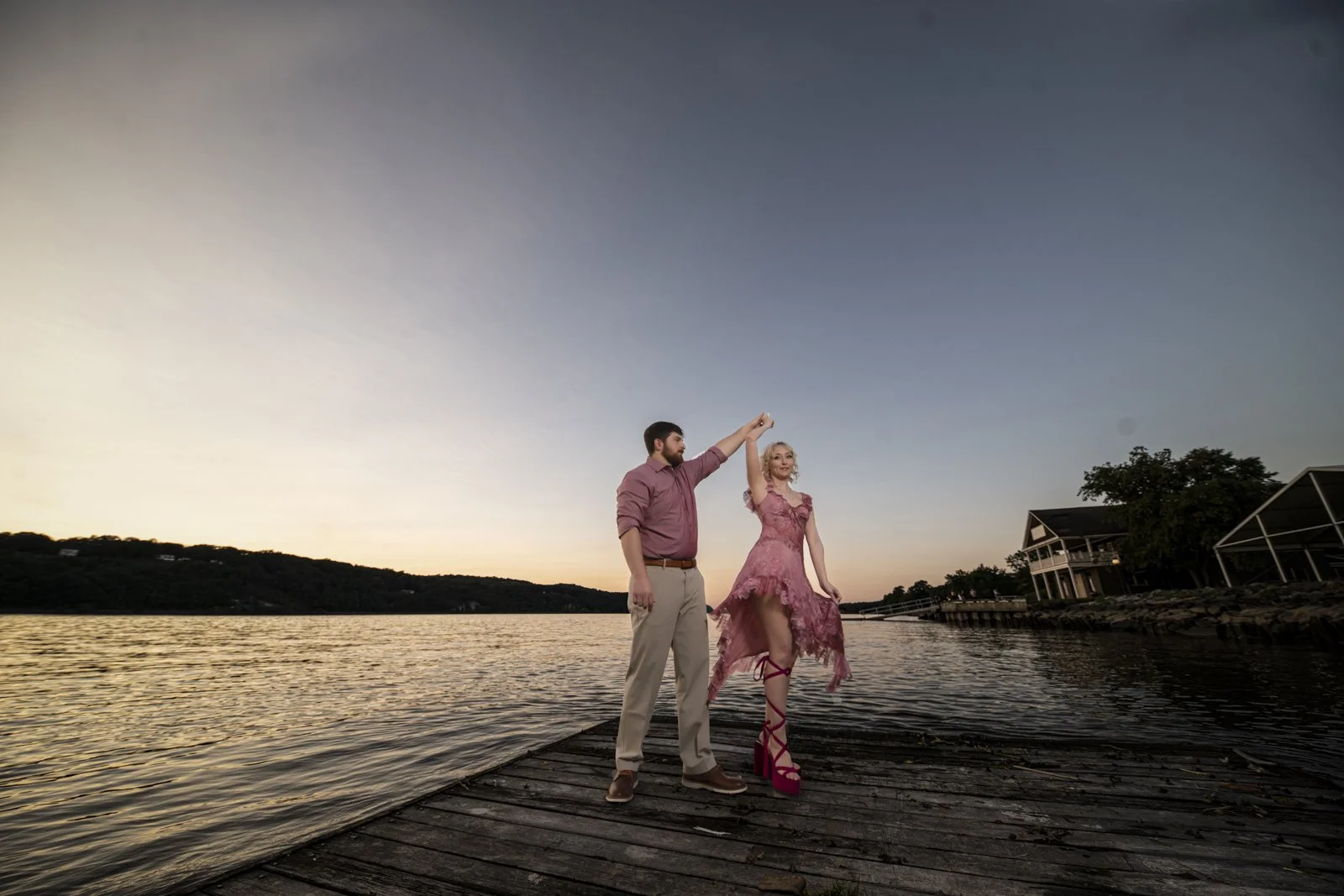 A man and woman dancing together on a wooden dock by a lake at sunset, with the man in a pink shirt and khakis, and the woman in a pink dress and pink high heels.
