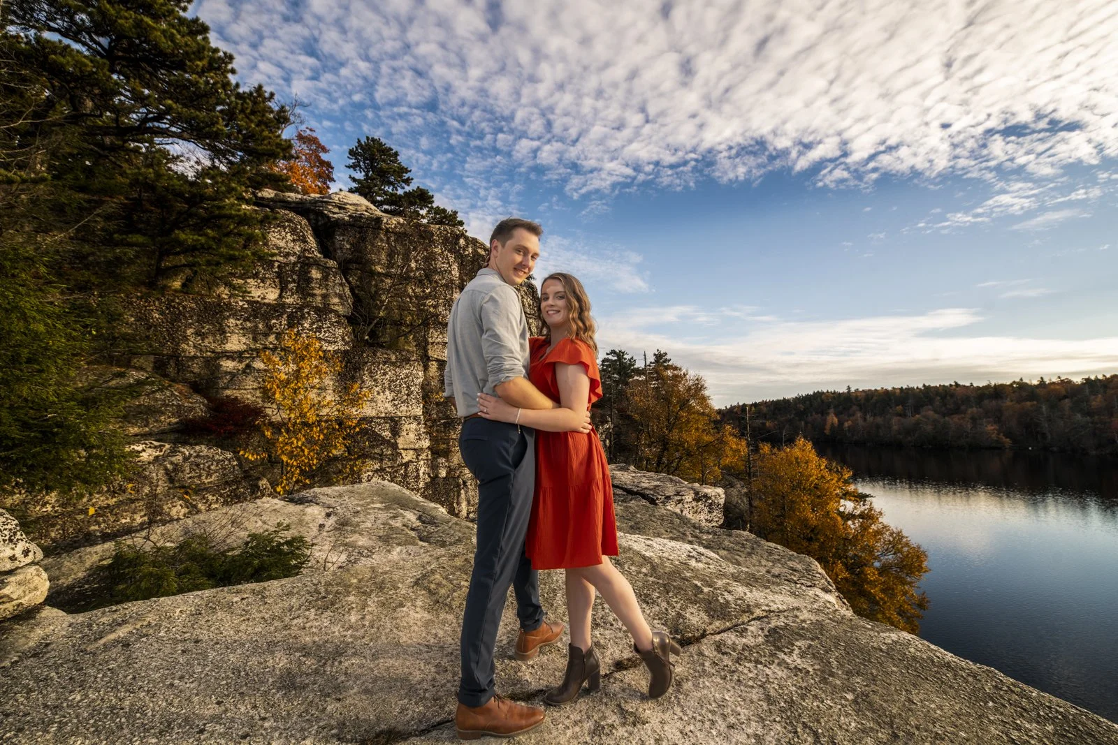 A couple stands on a rocky ledge near a body of water, surrounded by autumn trees, with a partly cloudy sky overhead.
