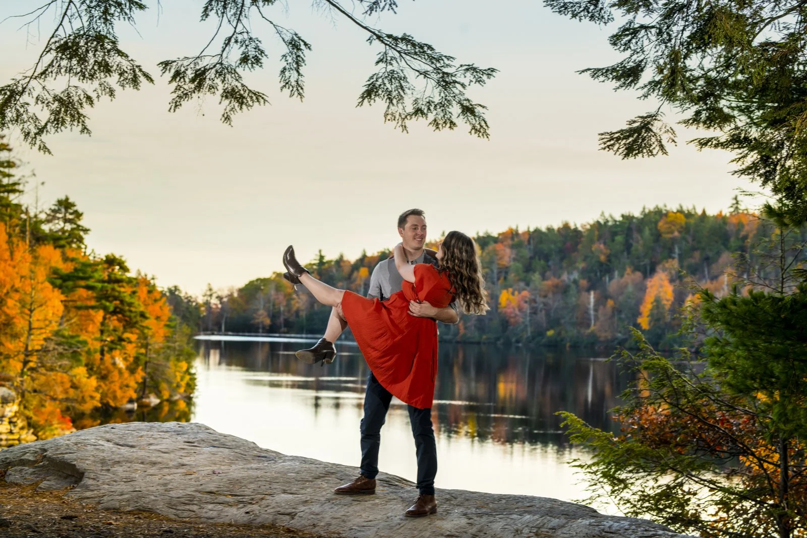A man is holding a woman in a red dress in his arms on a large rock beside a lake with autumn-colored trees in the background.