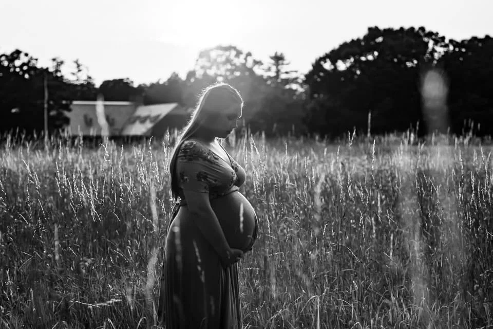 A pregnant woman standing in a field of tall grass, facing sideways with her hands on her belly, during dusk or dawn.