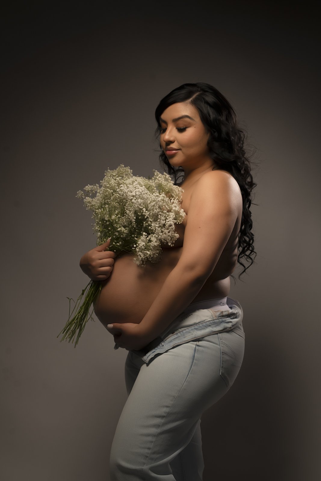 A pregnant woman with long, dark, curly hair holds a bouquet of white flowers while standing topless against a gray background.