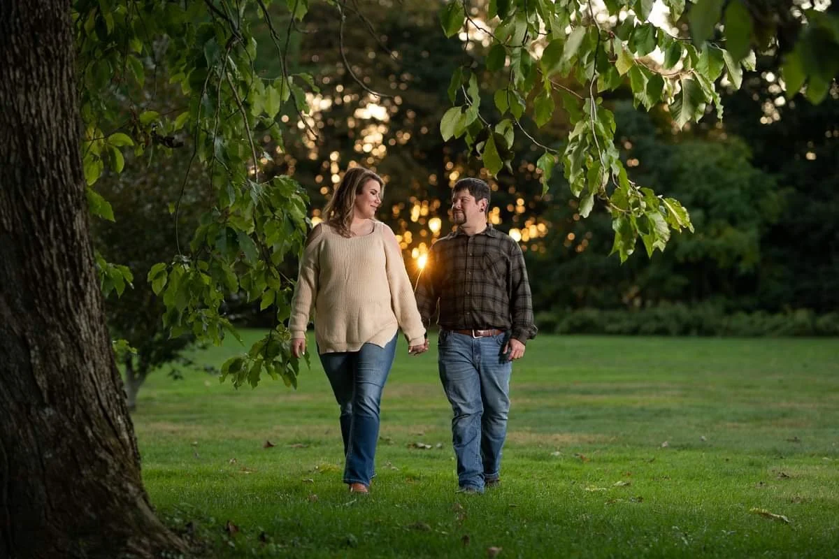 A couple walking hand in hand through a park at sunset, surrounded by green trees and grass.