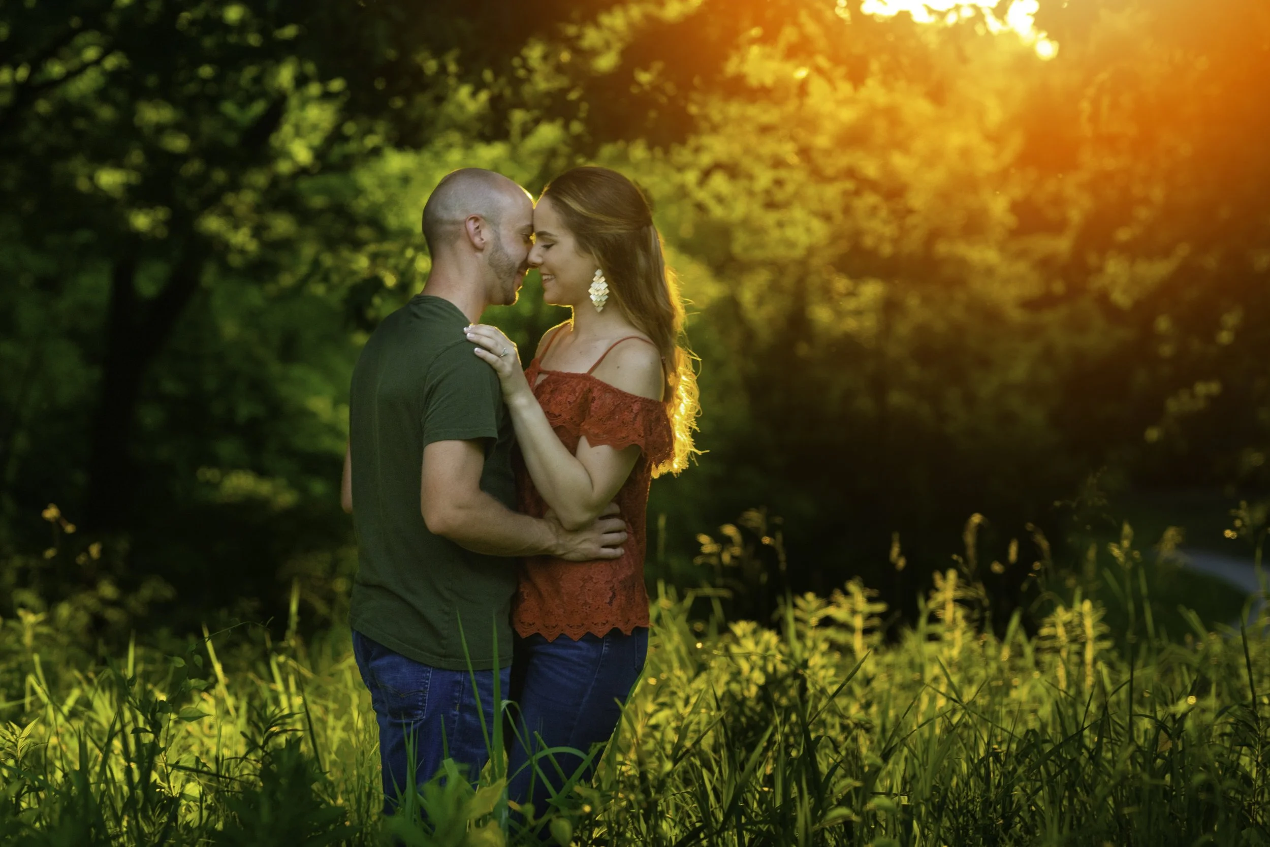 A couple in love standing close together in a green field during sunset, with their foreheads touching and smiling.