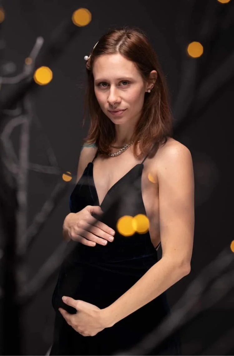 A young woman with shoulder-length brown hair, wearing a black spaghetti strap dress, holding a black clutch, posing in front of a dark background with yellow bokeh lights and branches in the foreground.