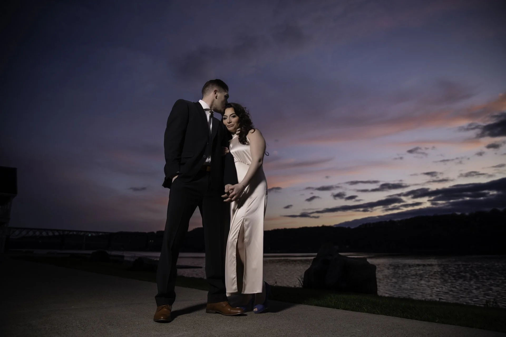 A couple dressed in formal attire standing on a waterfront promenade during sunset.