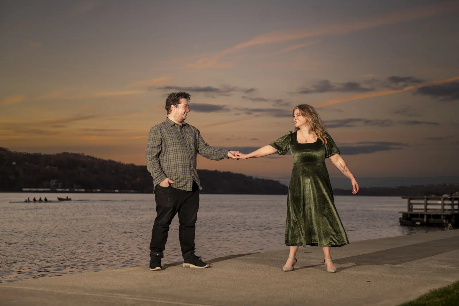 A couple stands on a waterfront promenade during sunset, holding hands and smiling at each other.