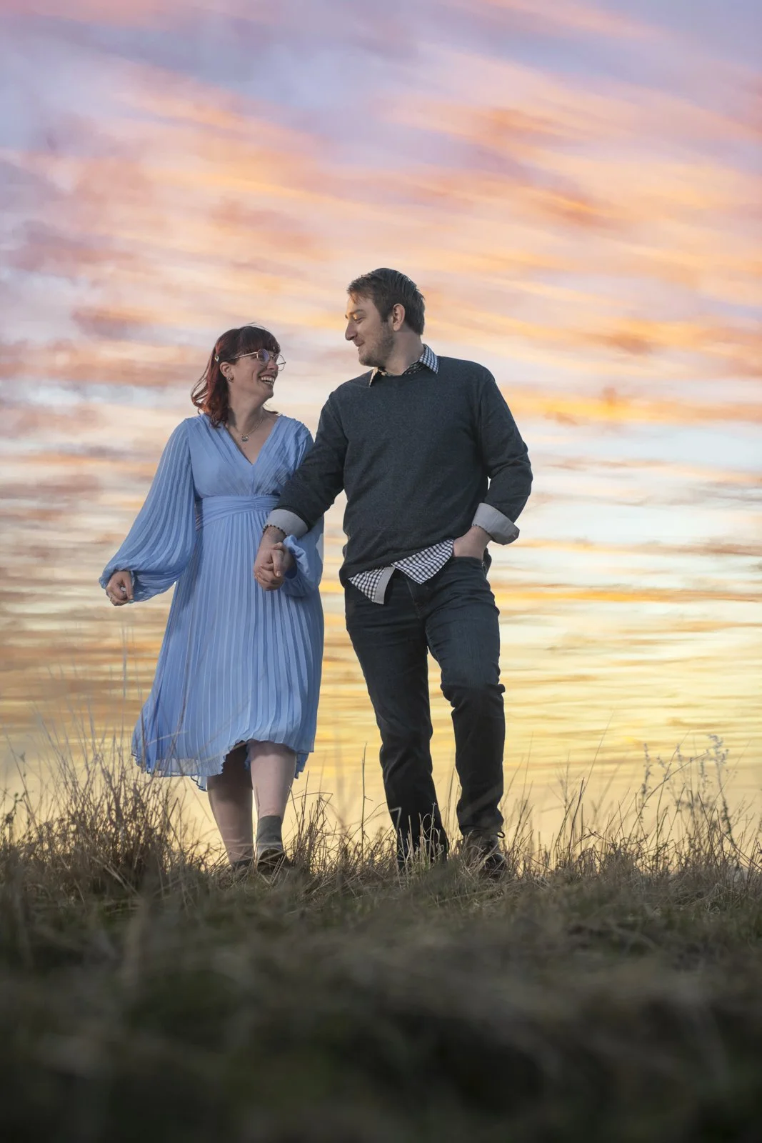 A couple walking hand in hand during sunset, smiling at each other.