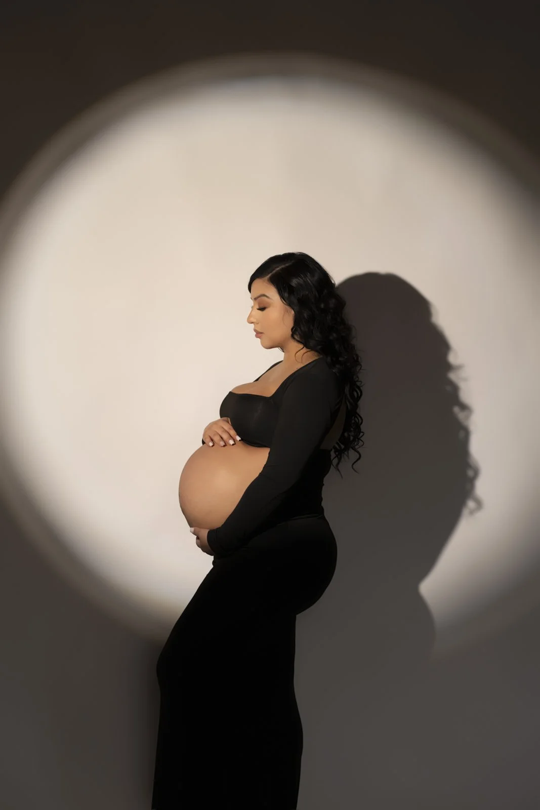 Pregnant woman with long black curly hair in black outfit standing in profile against a white background, shadow cast on wall.