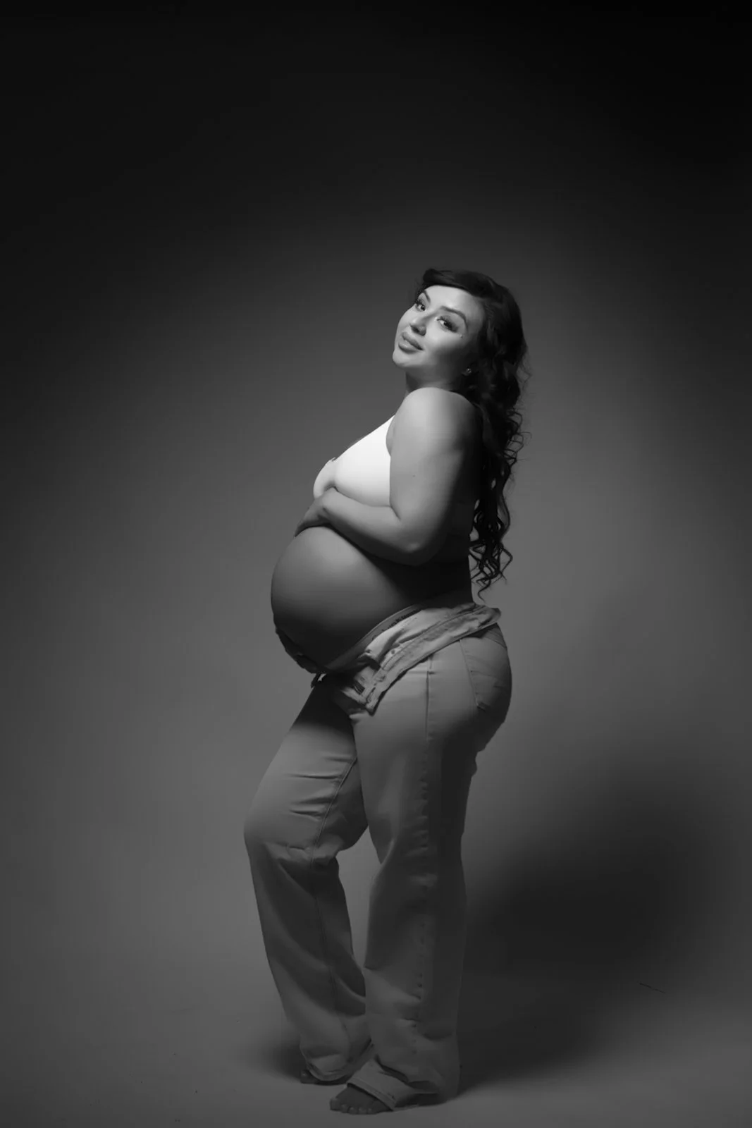 Black and white photo of a pregnant woman with long dark curly hair, wearing a white top and loose pants, standing against a gradient background.