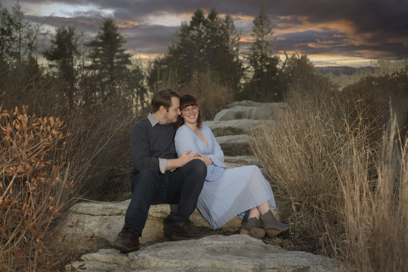 A man and woman sitting on rocks outdoors at sunset, embracing and smiling.