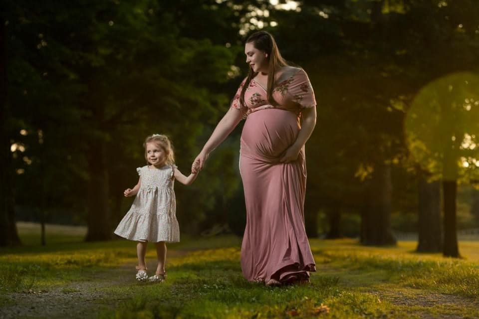 A pregnant woman in a pink dress holding hands with a young girl in a white dress walking outdoors in a park during sunset.