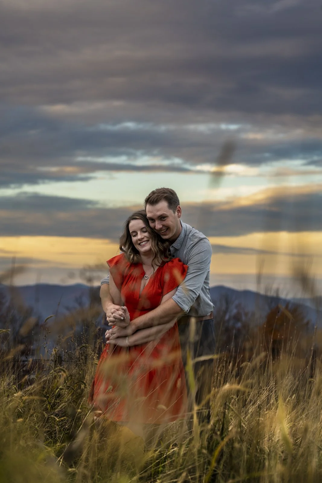 A happy couple embracing each other in a field with tall grass at sunset, with mountains in the background and a colorful cloudy sky.