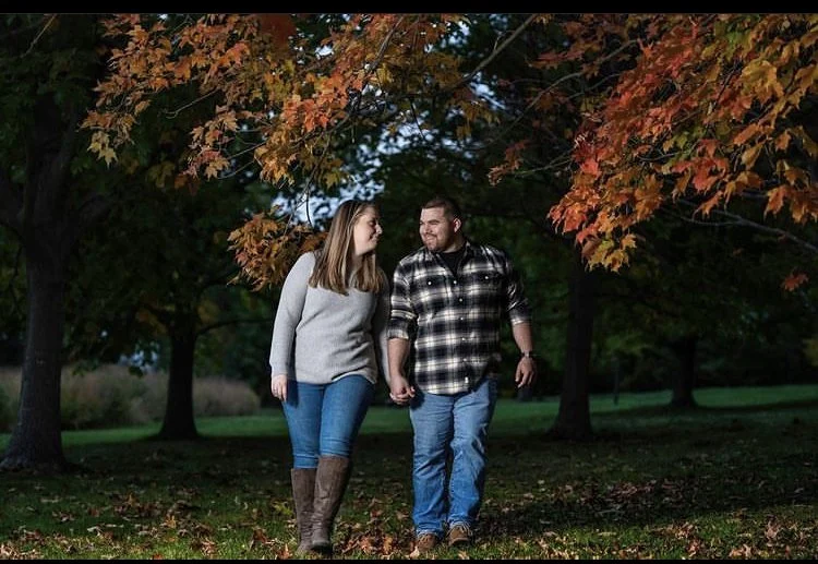A couple walking under orange autumn trees in a park, holding hands and smiling.