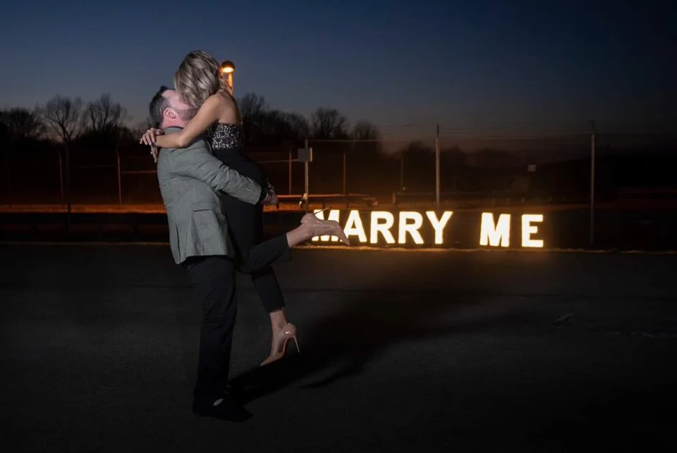 A man giving a woman a piggyback ride at night near a lit sign that says 'HARRY ME' on a dark street.