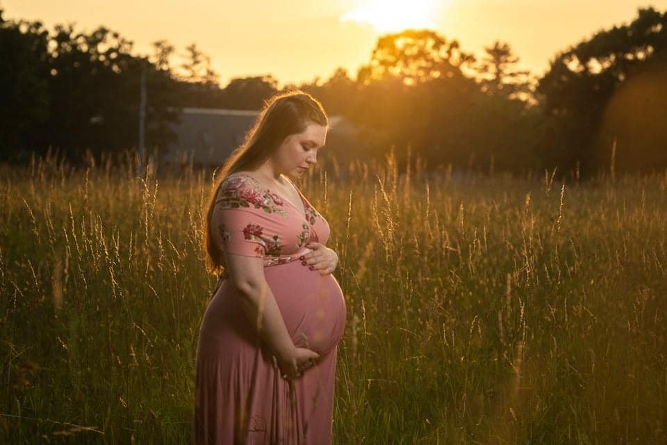 Pregnant woman in a pink floral dress standing in a field during sunset, gently holding her belly.