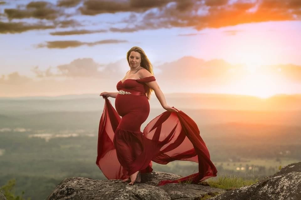 A pregnant woman in a red dress stands barefoot on a rocky hilltop during sunset, holding the flowing fabric of her dress. The sky is partly cloudy with a warm glow from the setting sun, and a vast landscape is visible in the background.