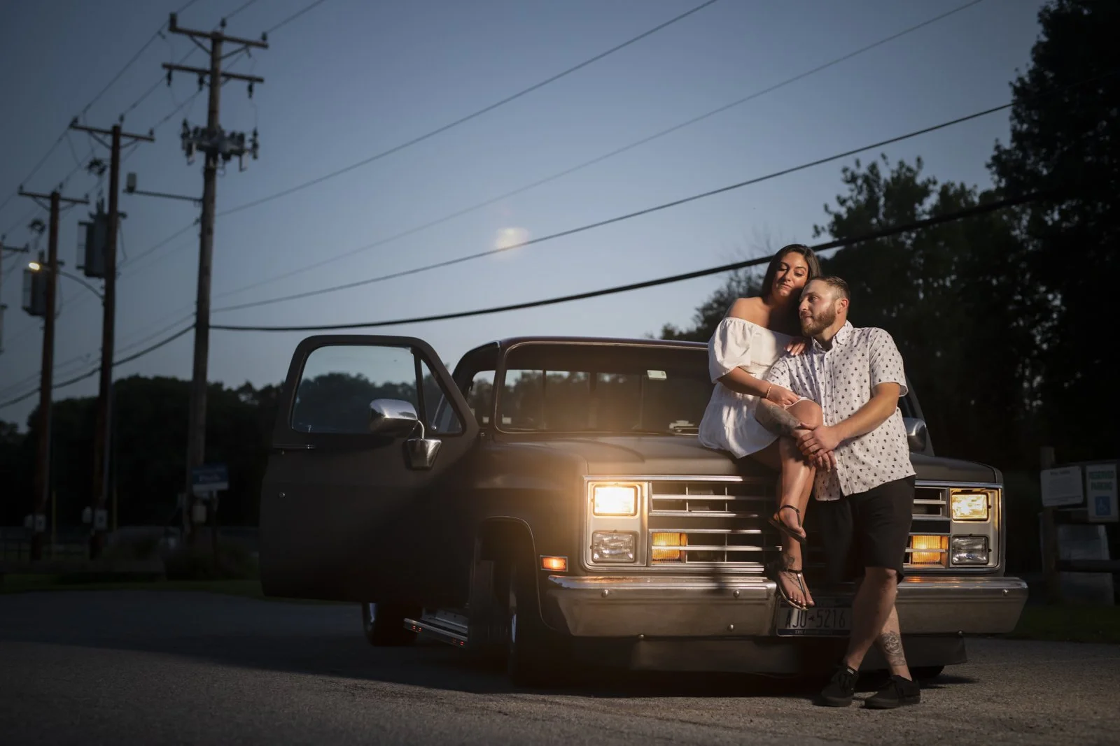 A couple sitting on a Cadillac car on the street at dusk, with power lines and trees in the background.