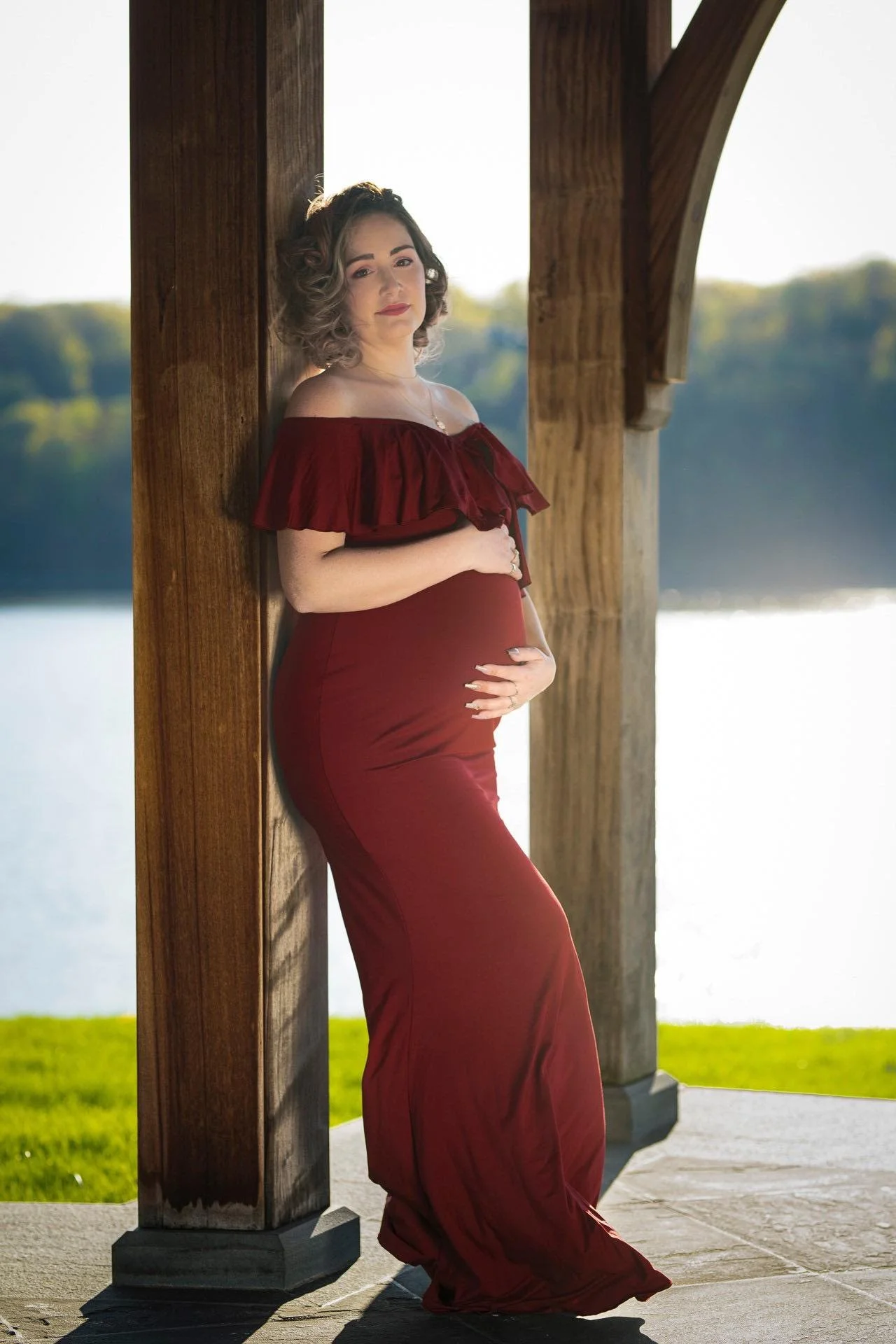 A pregnant woman in a red off-shoulder dress leaning against a wooden pillar outdoors near a body of water.