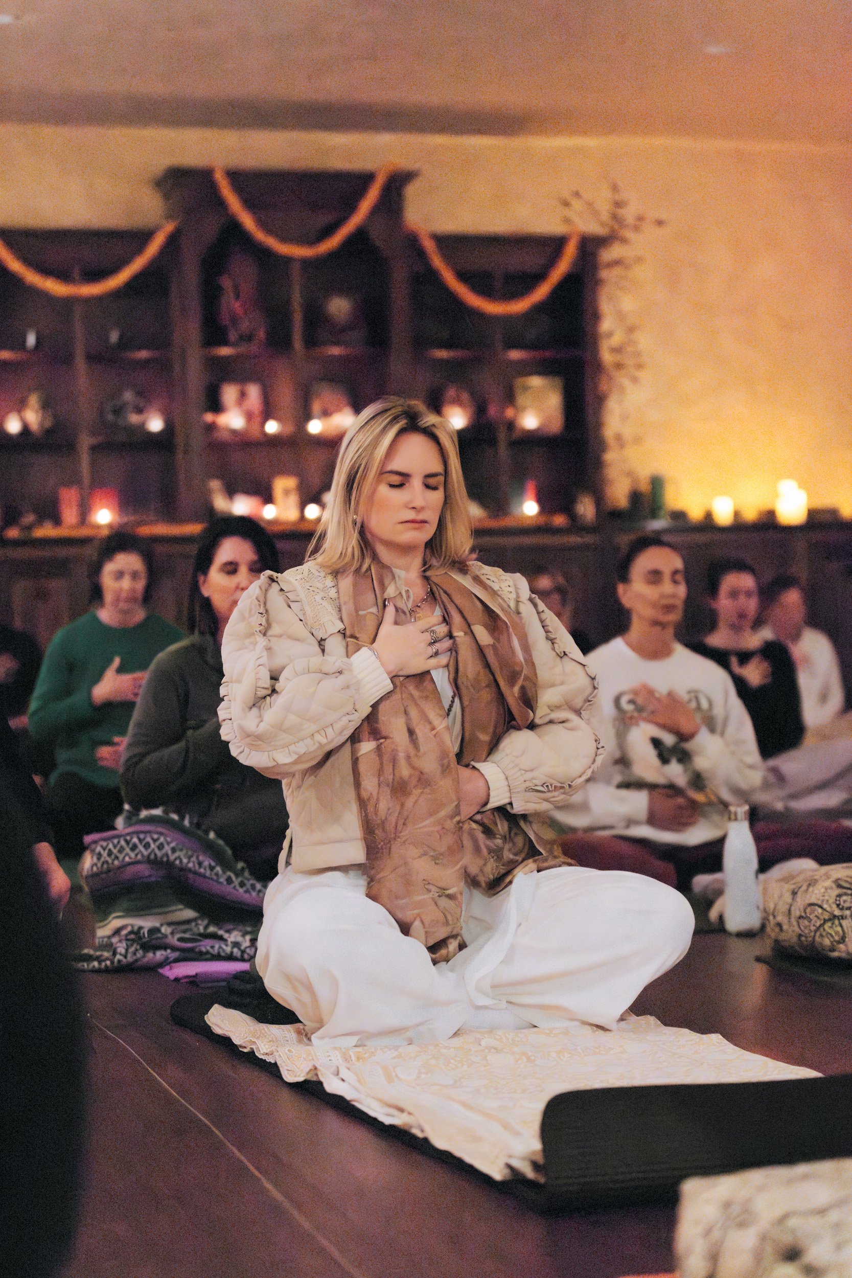 Group of women participating in a meditation or yoga class in a serene room with wooden shelves and spiritual decor.