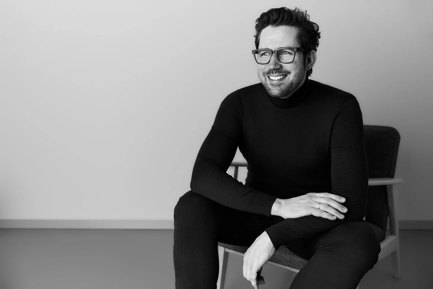 Ein Mann mit Brille sitzt mit dunkler Kleidung auf einem Sessel vor hellgrauem Hintergrund