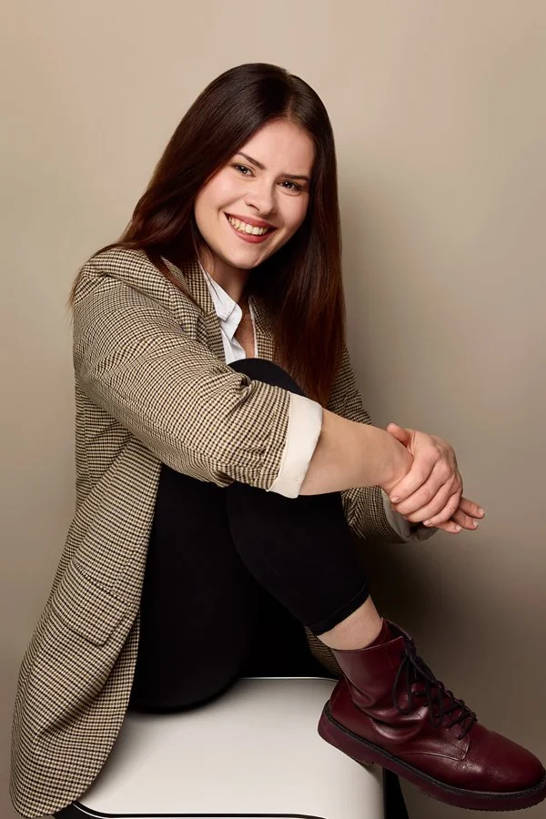 Ein Business Foto von einer Frau, die mit einem karierten Sakko auf einem weißen Barhocker sitzt und in die Kamera lächelt